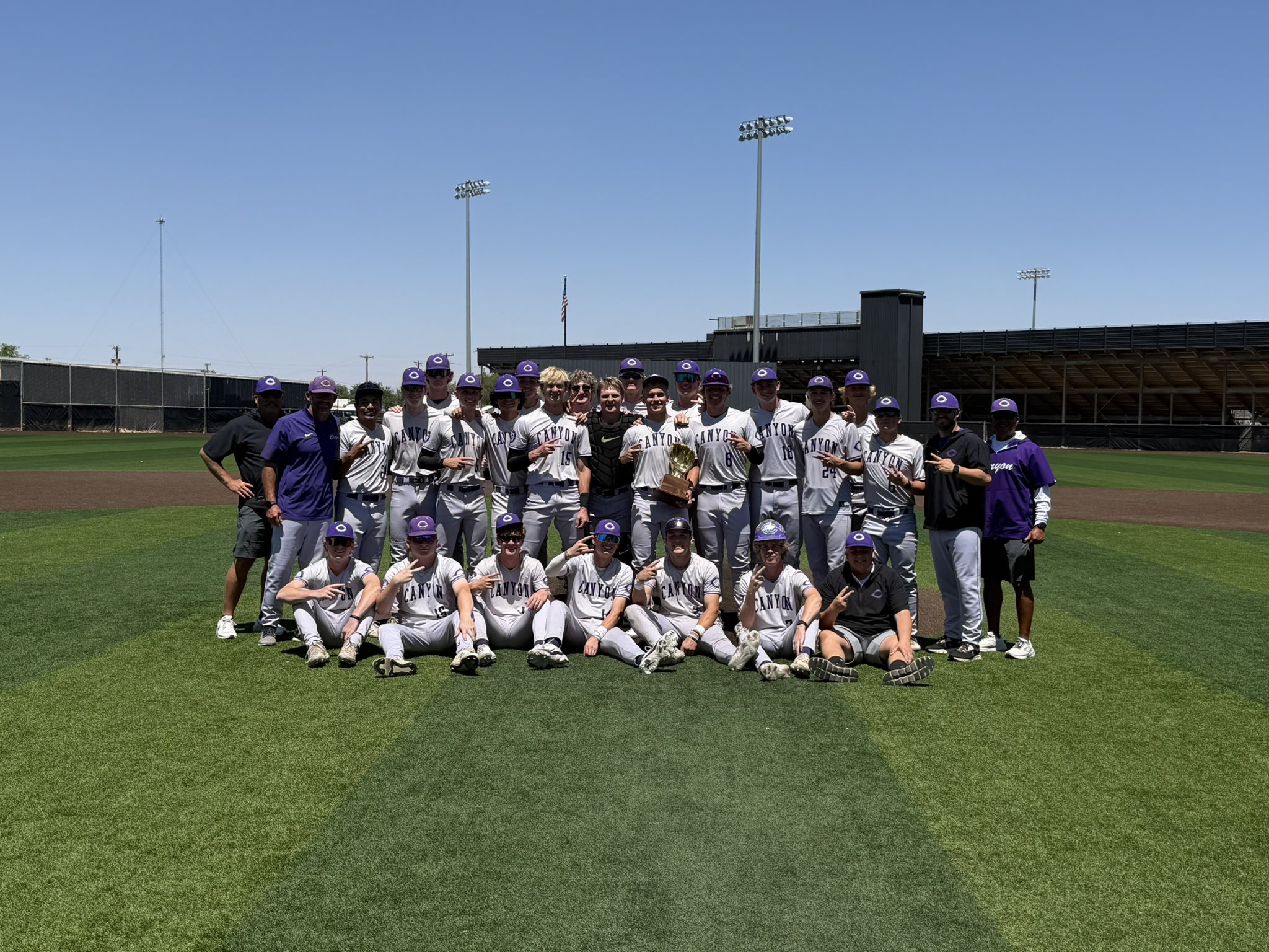 Baseball team in gray uniforms and purple hats posing with a trophy on the field.