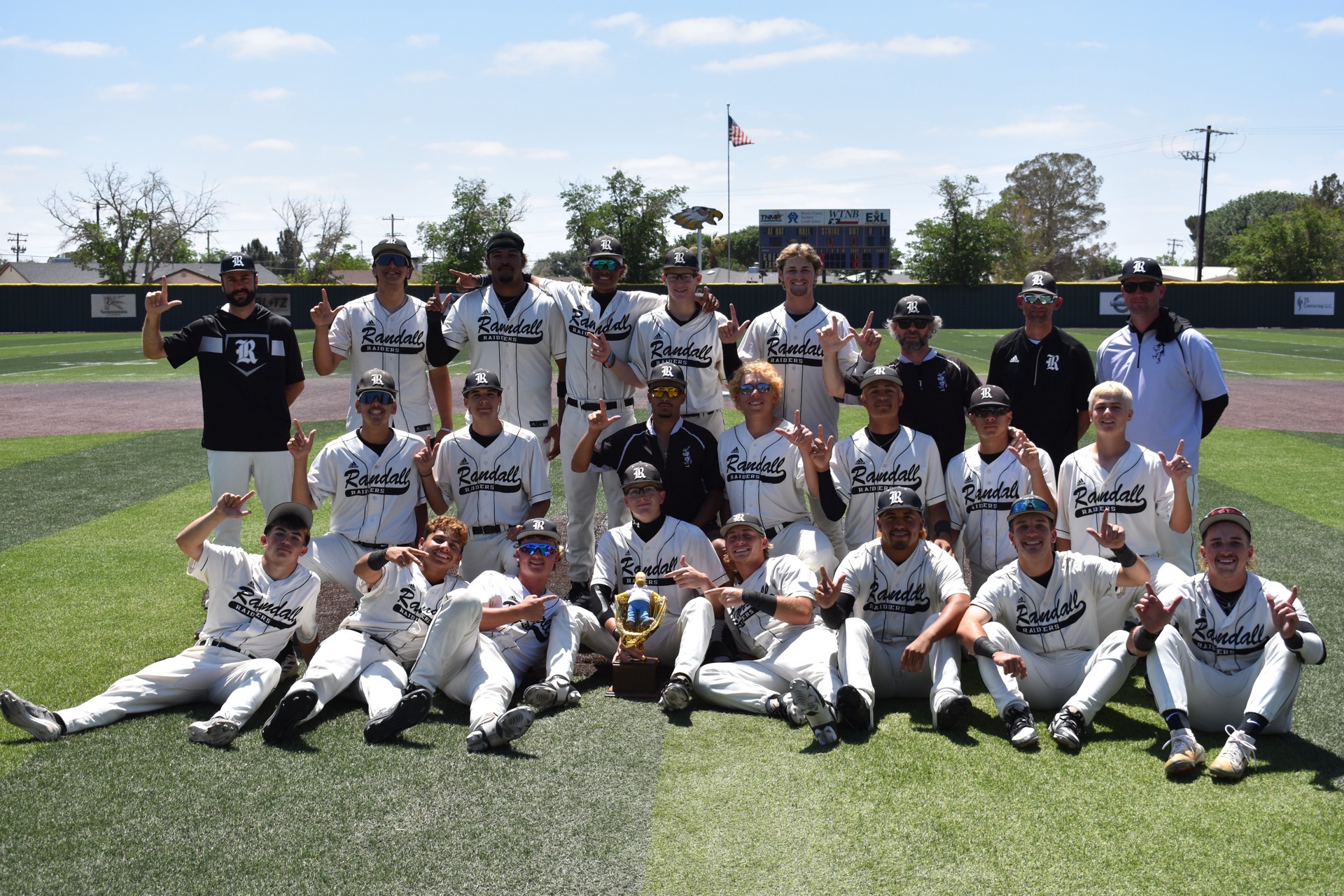 Smile-filled group photo of Randall Raiders baseball team posing on the field with a trophy at center and American flag in the background