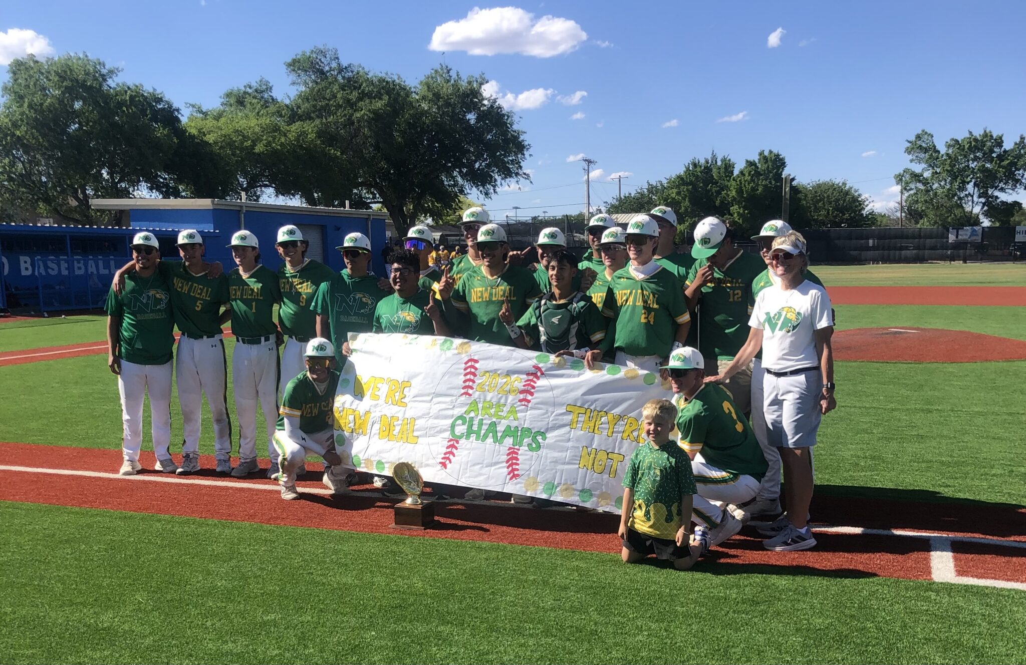 Youth baseball team in green jerseys posing with a banner and a small trophy on the field after a win, players wearing white helmets under a bright blue sky.