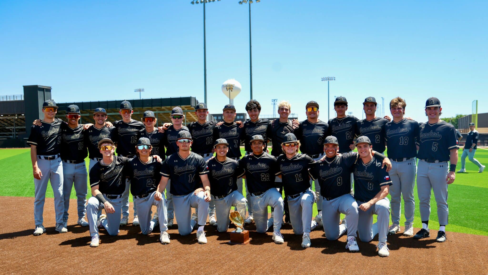 Group of Sandies baseball players posing for a team photo on the field with a trophy in front of them.