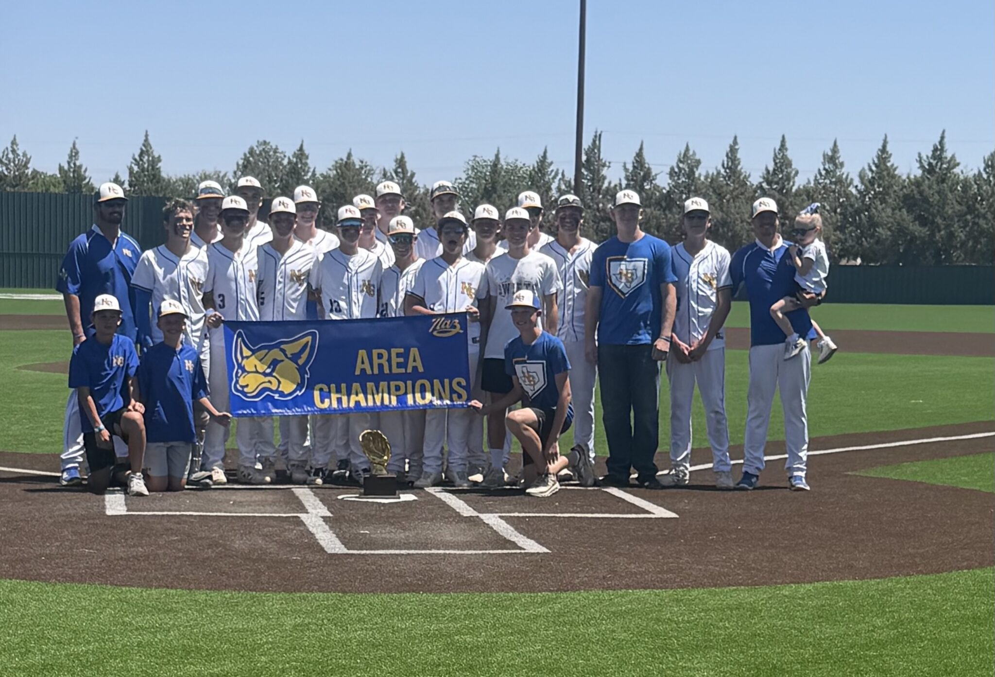 Group of baseball players in white uniforms and caps posing on the field with a blue banner reading 'AREA CHAMPIONS' and a trophy in front. Coaches and a child are present.