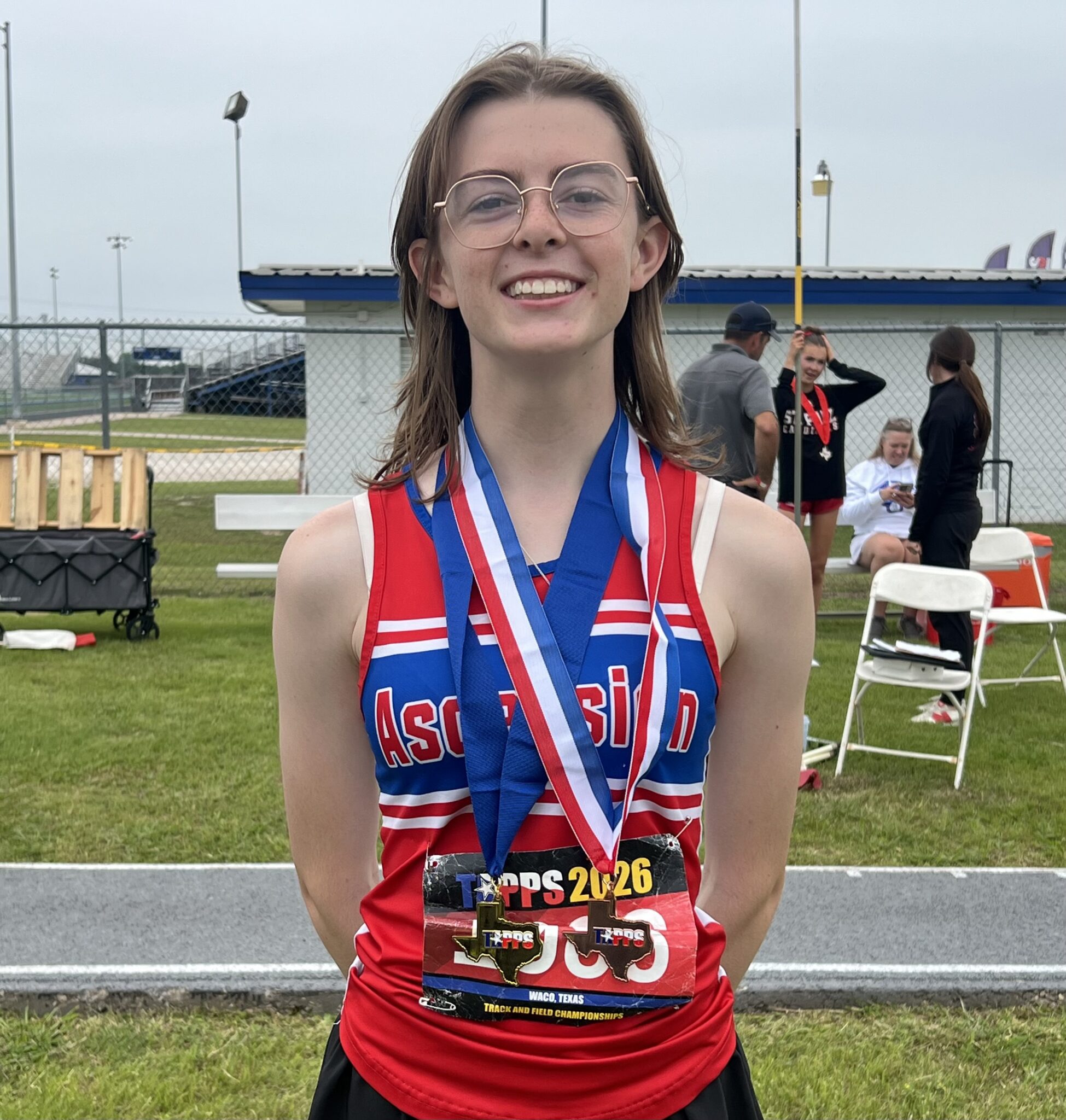 Smiling young female track athlete in red and blue uniform with medals around her neck on a grassy field after a race.