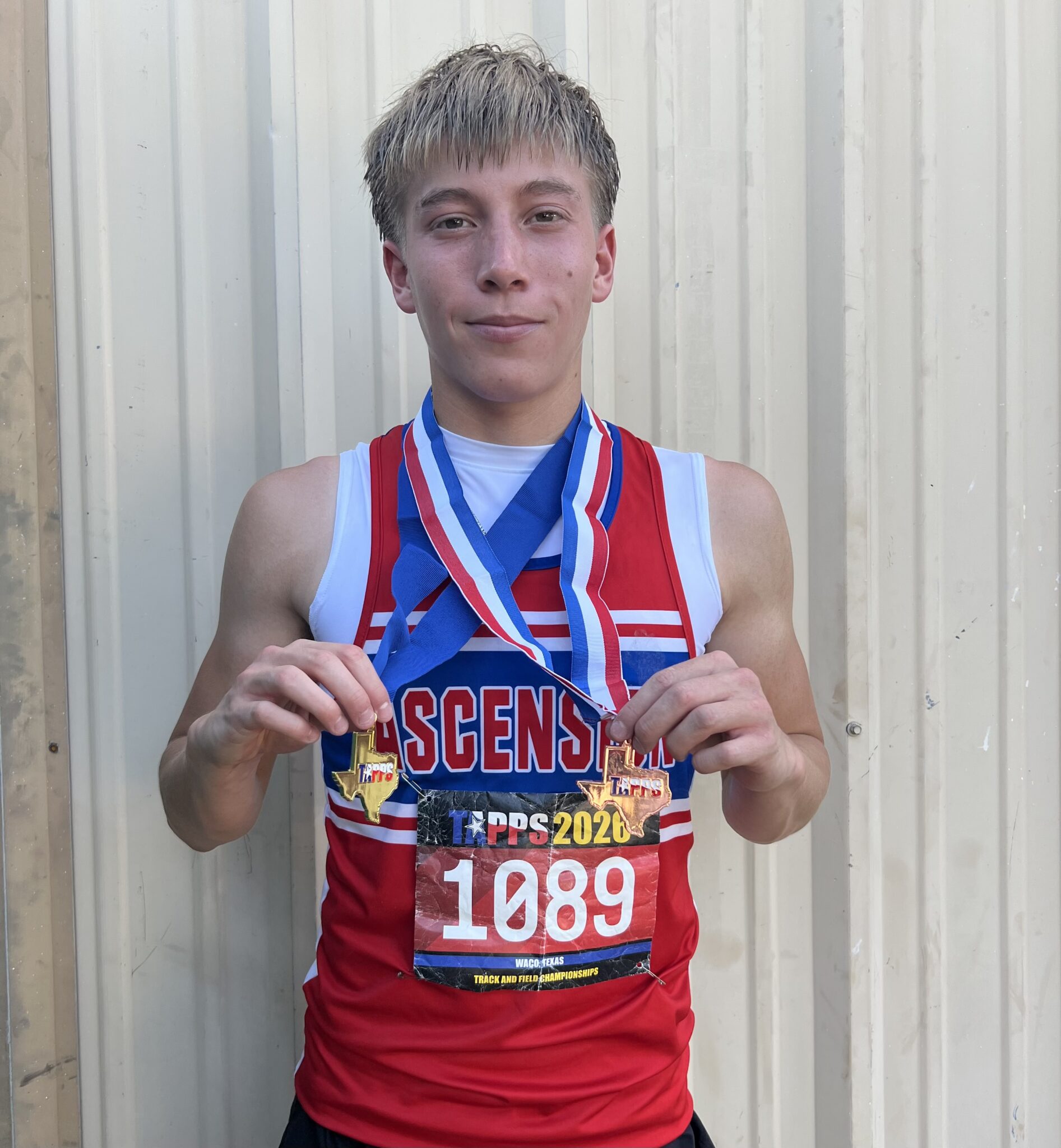 Young runner with multiple medals on red, white, and blue ribbons, posing in front of a metal backdrop.