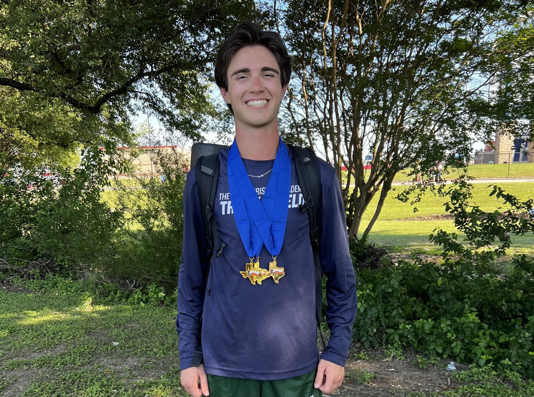 Smiling young man outdoors wearing a blue ribbon medal necklace and a backpack, with trees in the background.