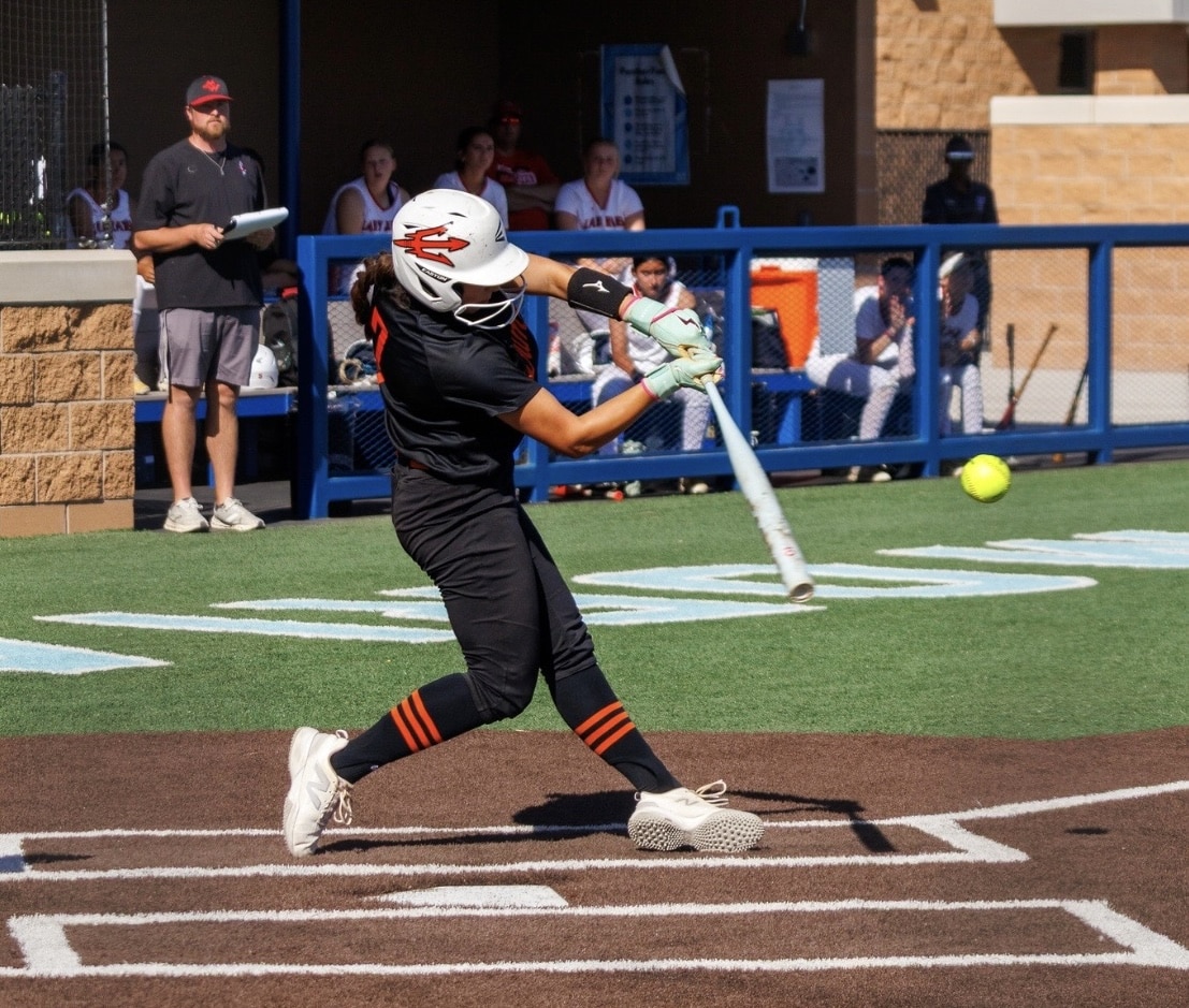 Female softball batter in black uniform with orange stripes swings at a pitch near home plate; yellow ball in play behind her.