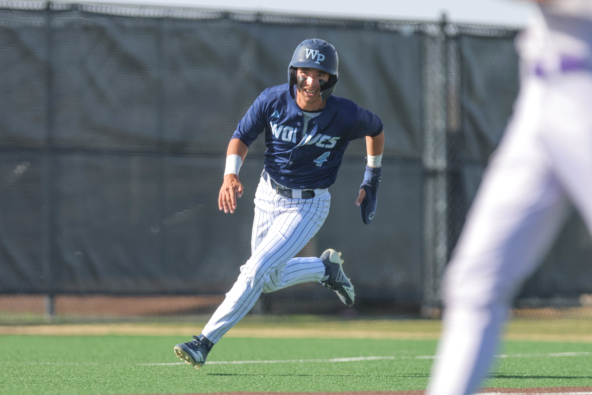 Baseball player in blue uniform running on the field, helmet on, with a blurred figure in foreground on the right.