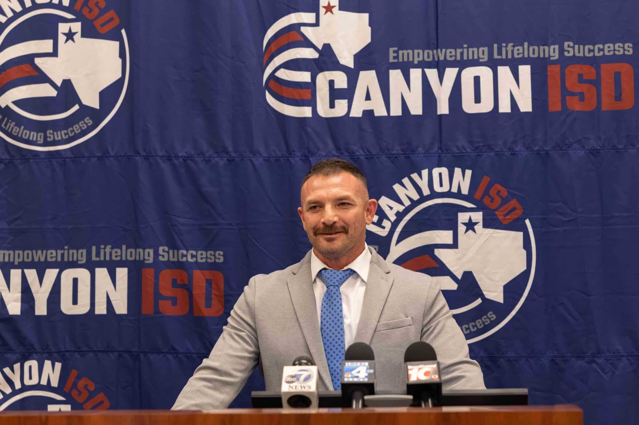 WPHS_Coach-7 | Press Pass Sports Man in a gray suit and blue tie at a press conference with microphones on the table, backdrop reads Canyon ISD logo and slogan.