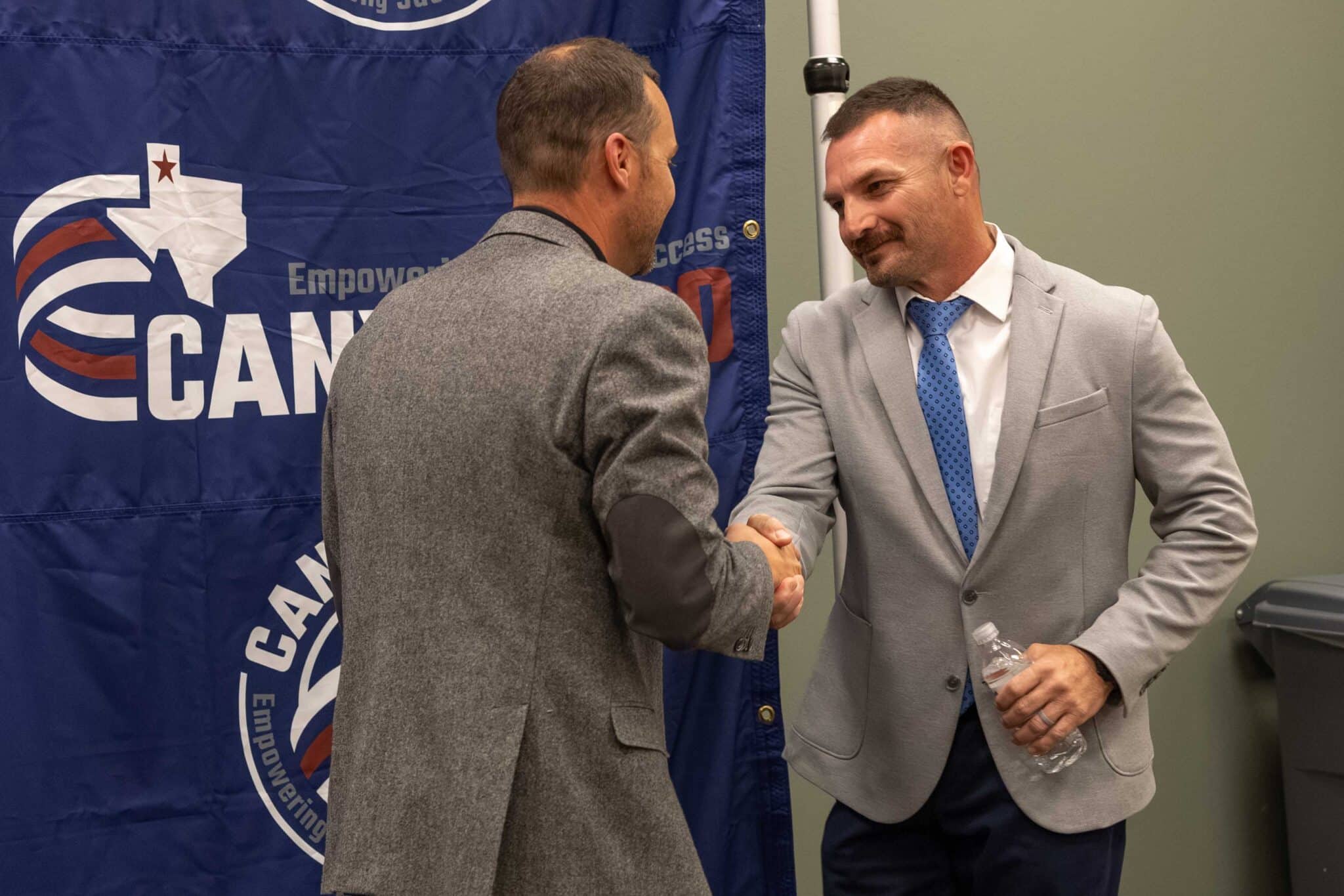 WPHS_Coach-5 | Press Pass Sports Two businessmen shake hands in front of a blue banner with a circular logo behind them.