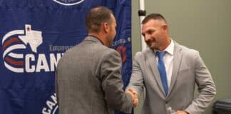 Two businessmen shake hands in front of a blue banner with a circular logo behind them.