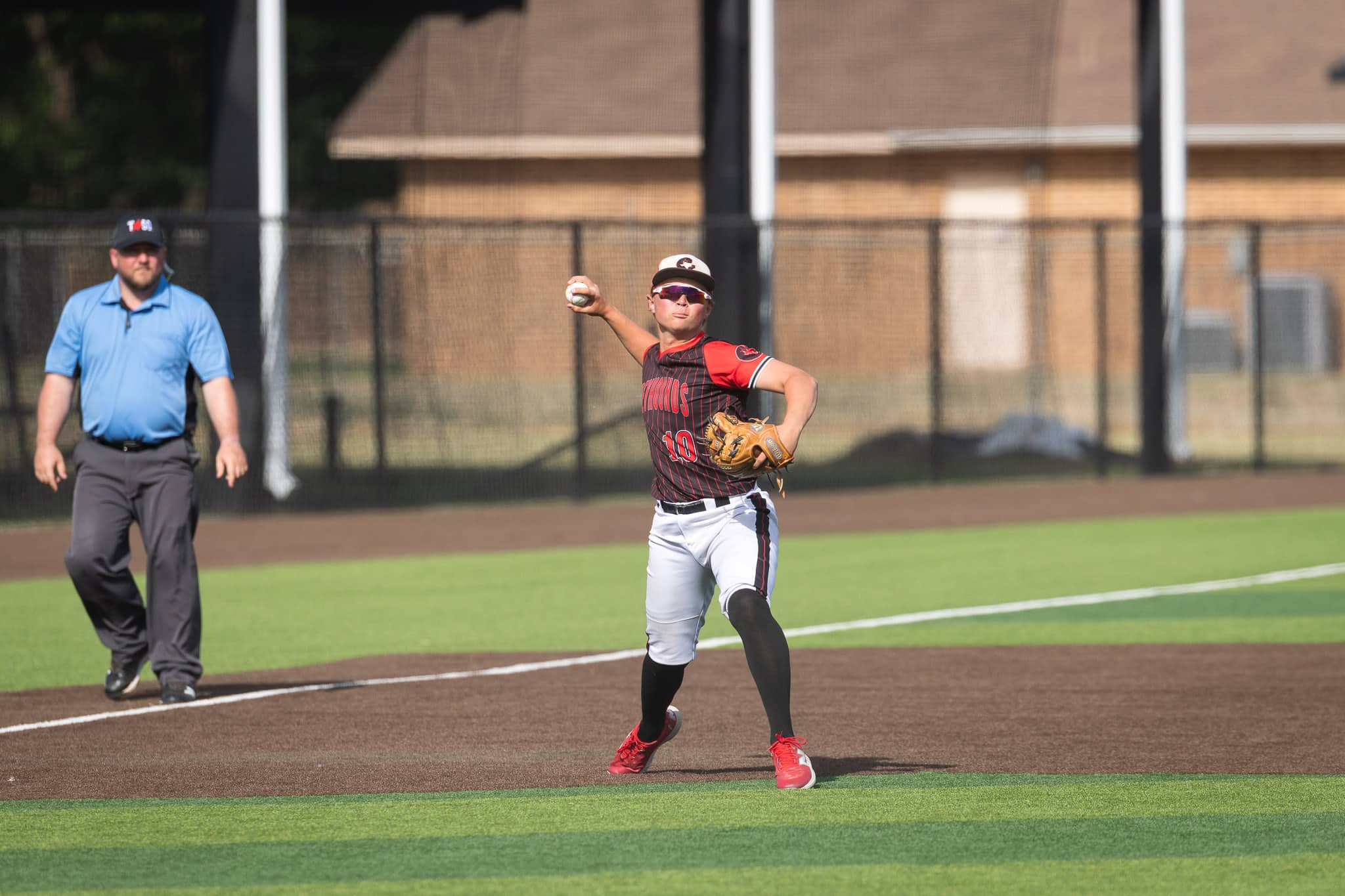 Baseball pitcher in red and black uniform mid-throw on the mound, with an official in a blue shirt watching from the left.