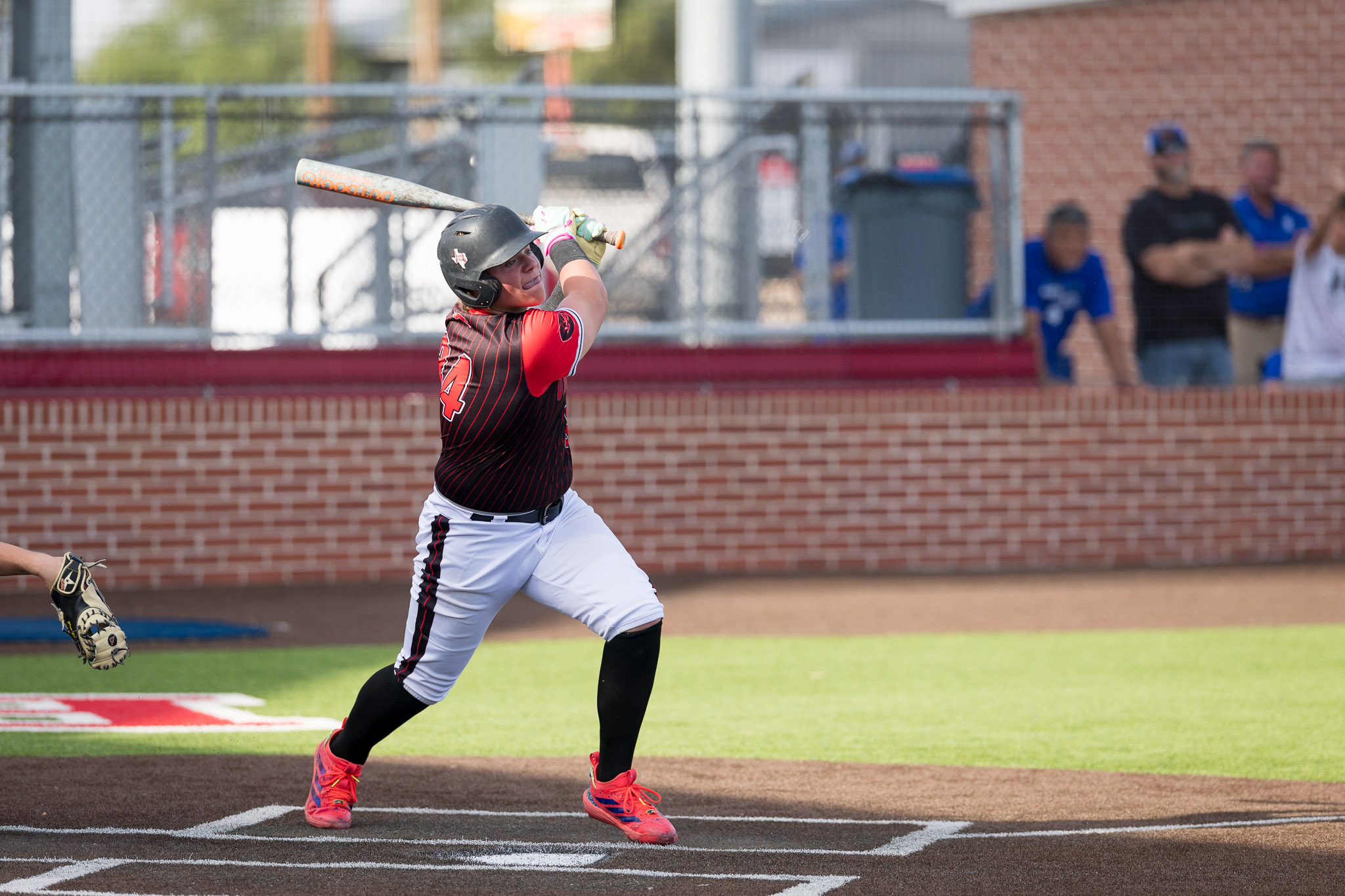 Female softball player in red-and-black uniform swings a bat at a pitch on a ballfield, helmet on, with a brick backstop behind.
