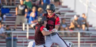 Two baseball players in black-and-red uniforms celebrate midair, embracing after a play, helmets on, stadium stands blurred behind them.