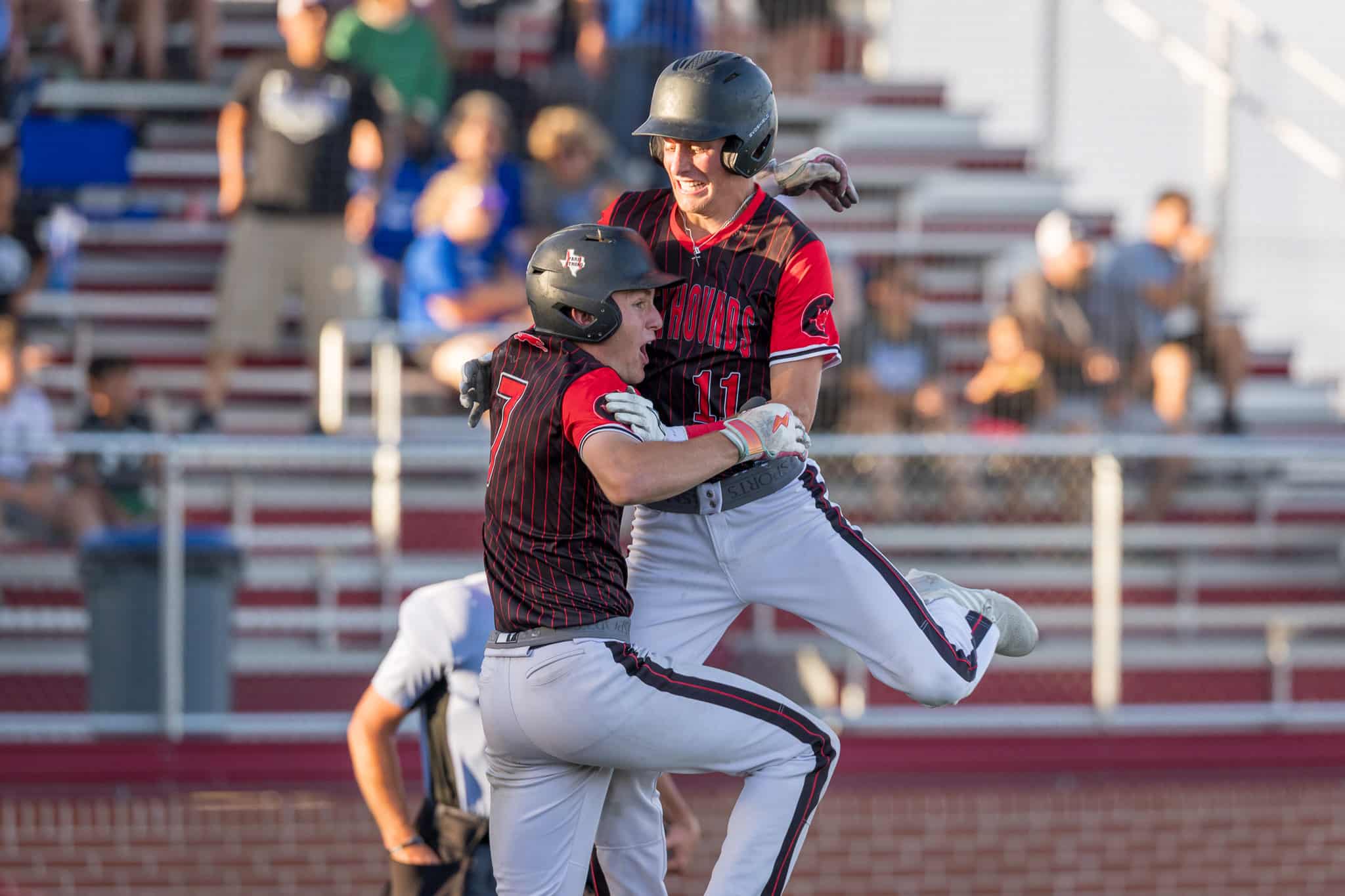 Two baseball players in black-and-red uniforms celebrate midair, embracing after a play, helmets on, stadium stands blurred behind them.