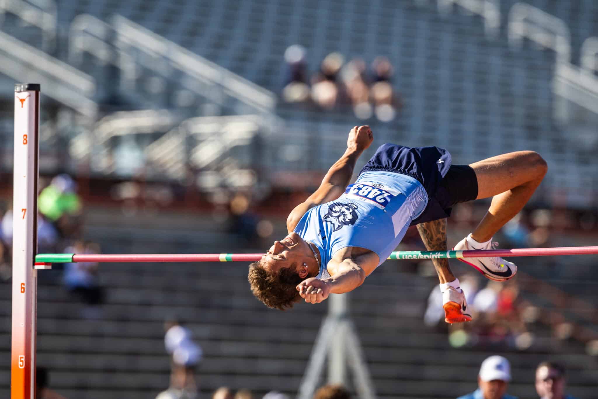 100A2226 | Press Pass Sports Athlete in a blue uniform clears a high jump bar in a stadium setting, mid-air.