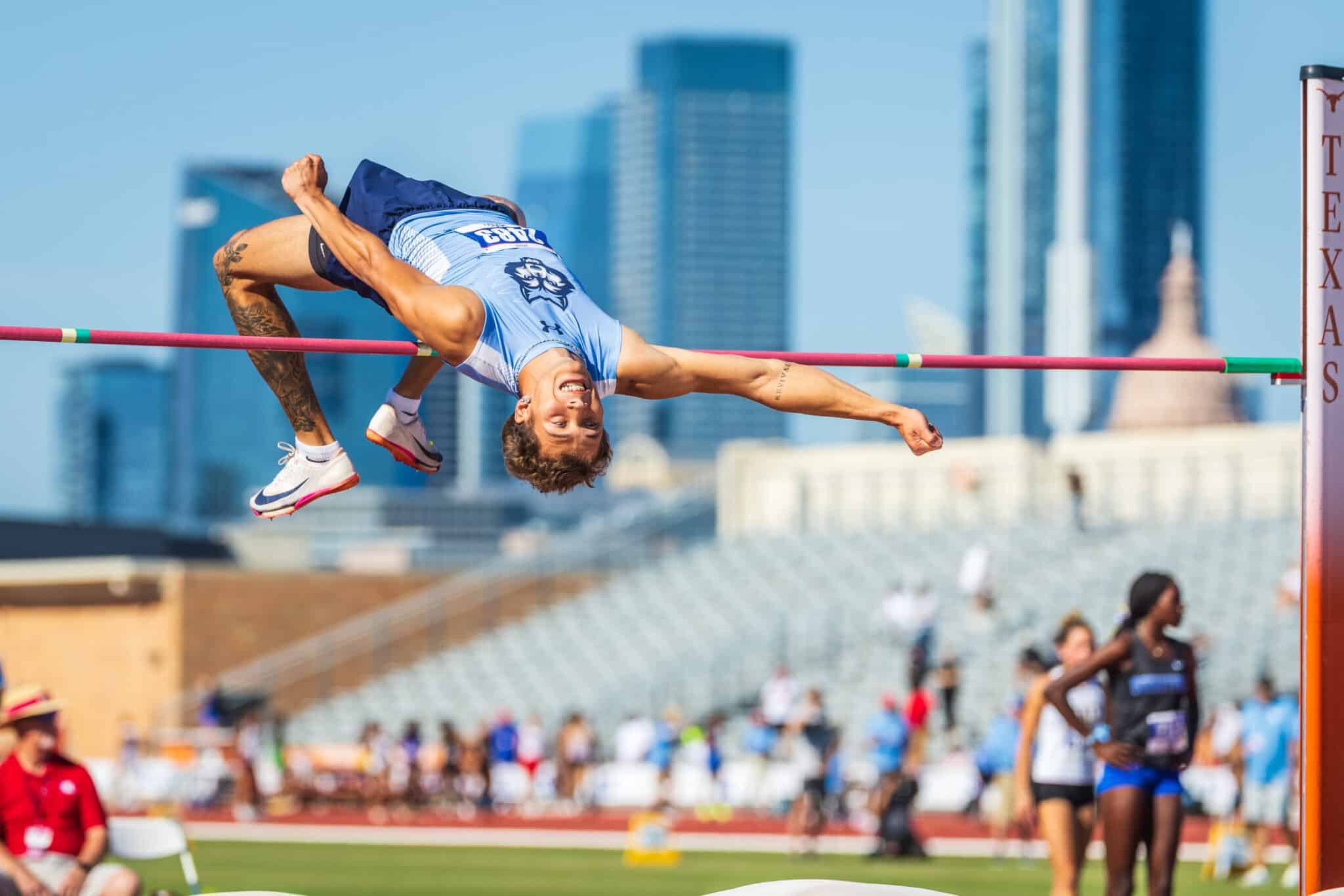 100A2366 | Press Pass Sports Male high jumper in a light blue uniform clears a pink bar in a stadium with a city skyline behind him.