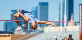Male high jumper in a light blue uniform clears a pink bar in a stadium with a city skyline behind him.