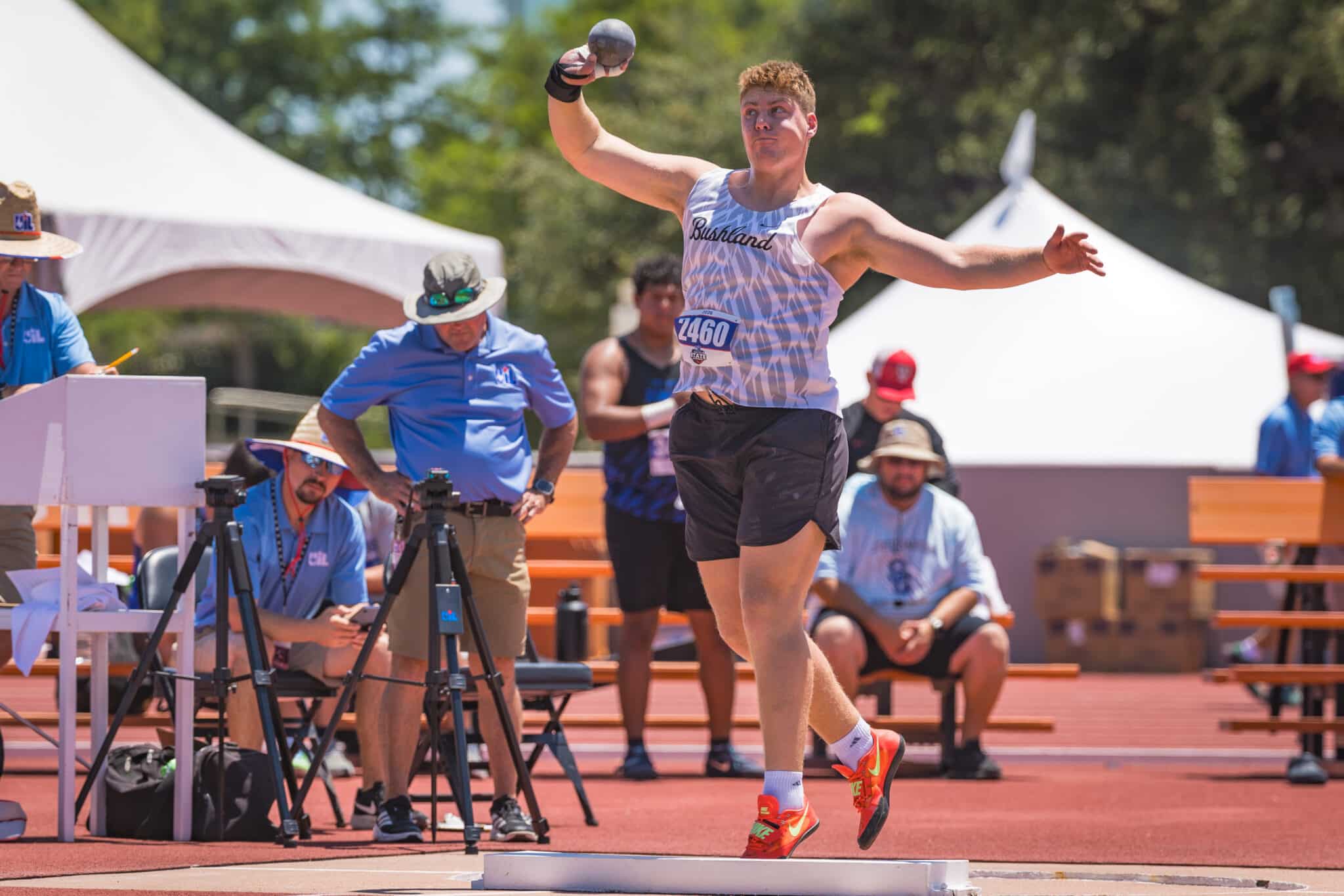 100A3695 | Press Pass Sports Male shot-put athlete mid-throw during a track meet, wearing a white patterned singlet and red sneakers, with officials and tents in the background.