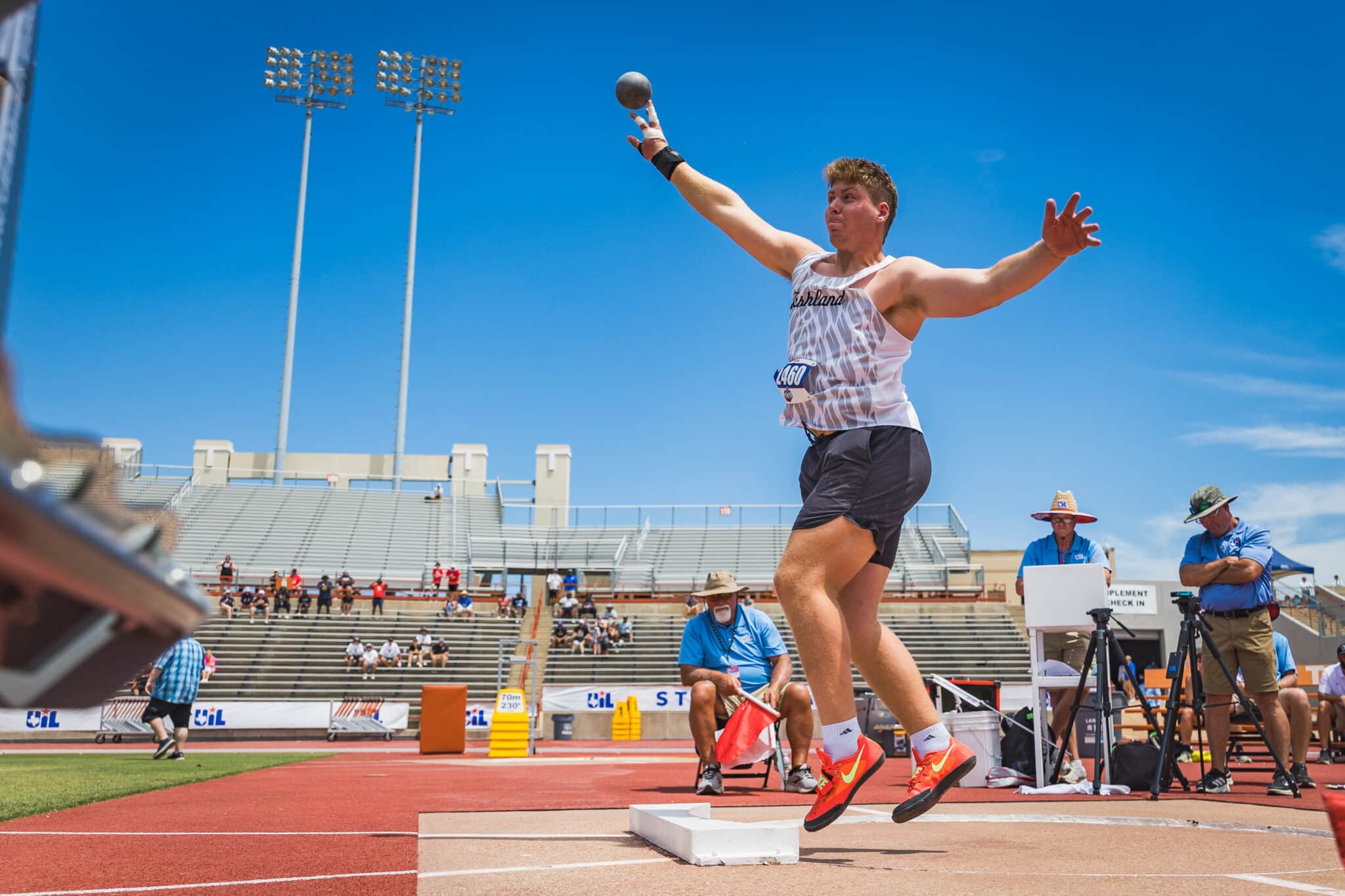 DE3_8621 | Press Pass Sports Male track and field athlete in a white singlet gliding mid‑air during a shot put event at an outdoor stadium, with spectators in the stands under a blue sky.