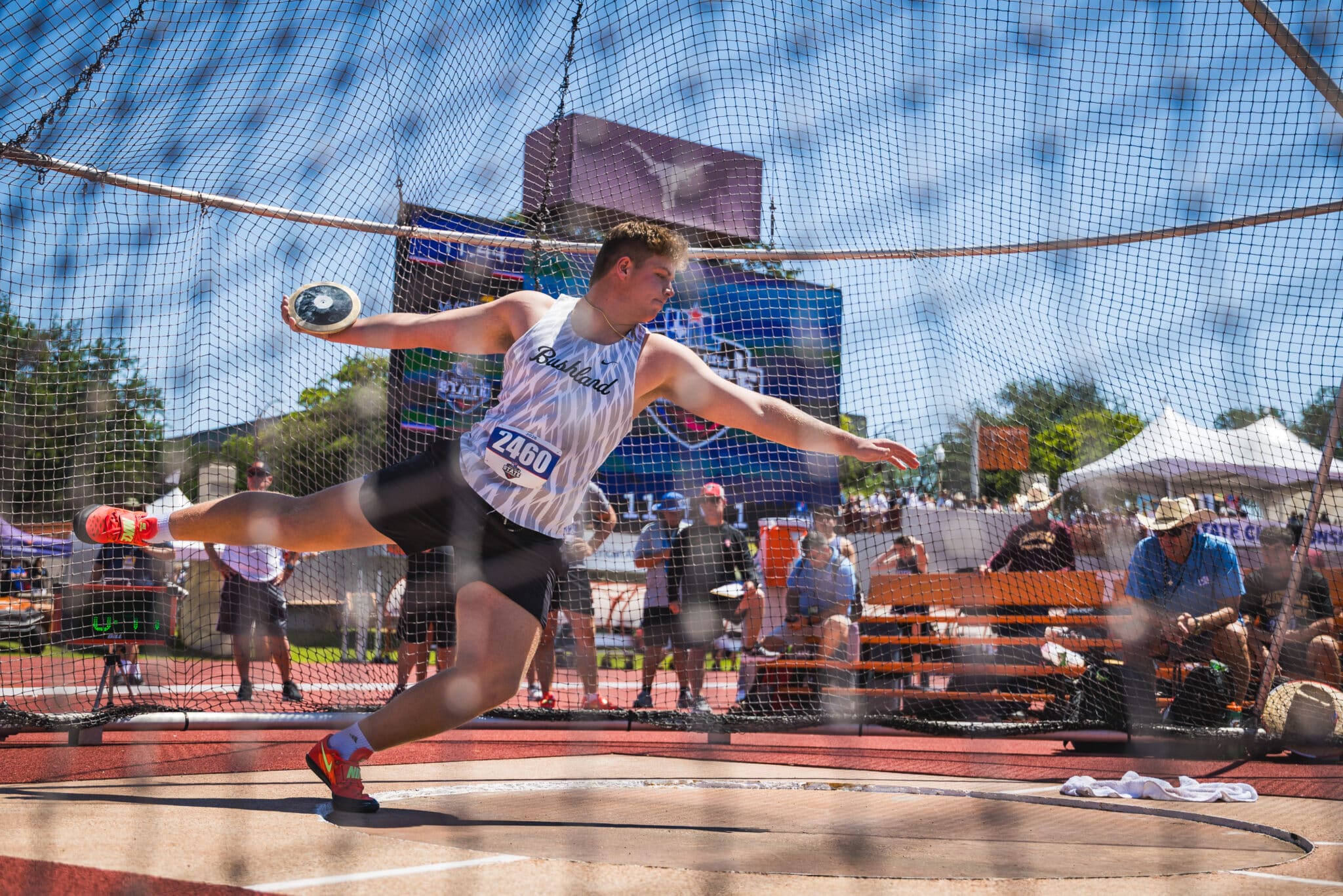DE3_8491A | Press Pass Sports Athlete in mid-throw on a track field, disc in hand, net enclosure around the throwing circle, crowd watching in the background.