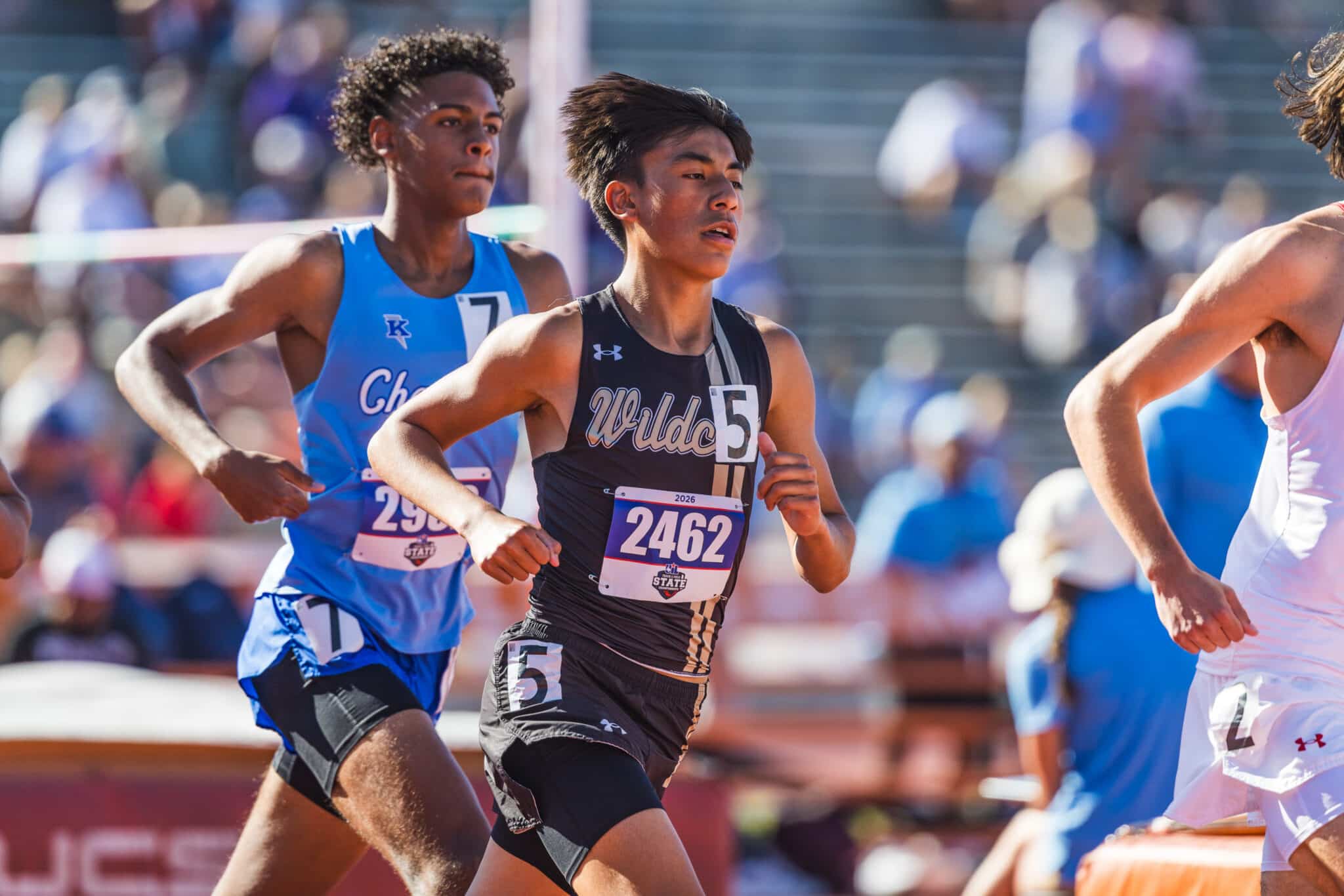 Three male runners sprinting on a track, center athlete in black with bib 2462, flanked by blue- and white-jersey runners.