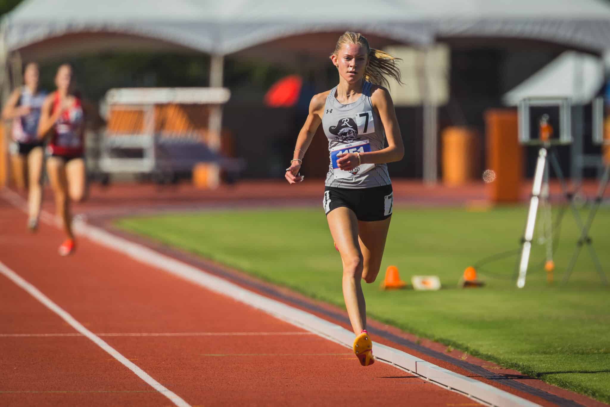 Female athlete sprinting on a red track, gray sleeveless top and black shorts, bib number visible in foreground with determined expression.