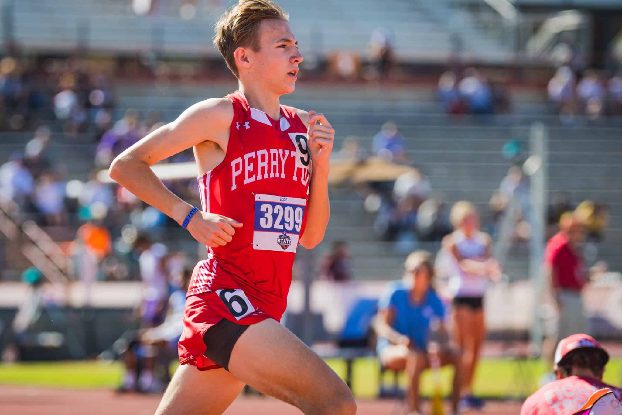 Teenage male runner in red Perry jersey mid-race on track with crowds in background, bib 3299 visible on chest.