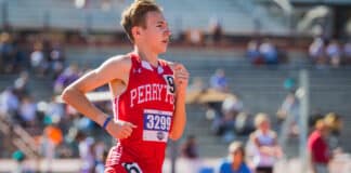 Teenage male runner in red Perry jersey mid-race on track with crowds in background, bib 3299 visible on chest.