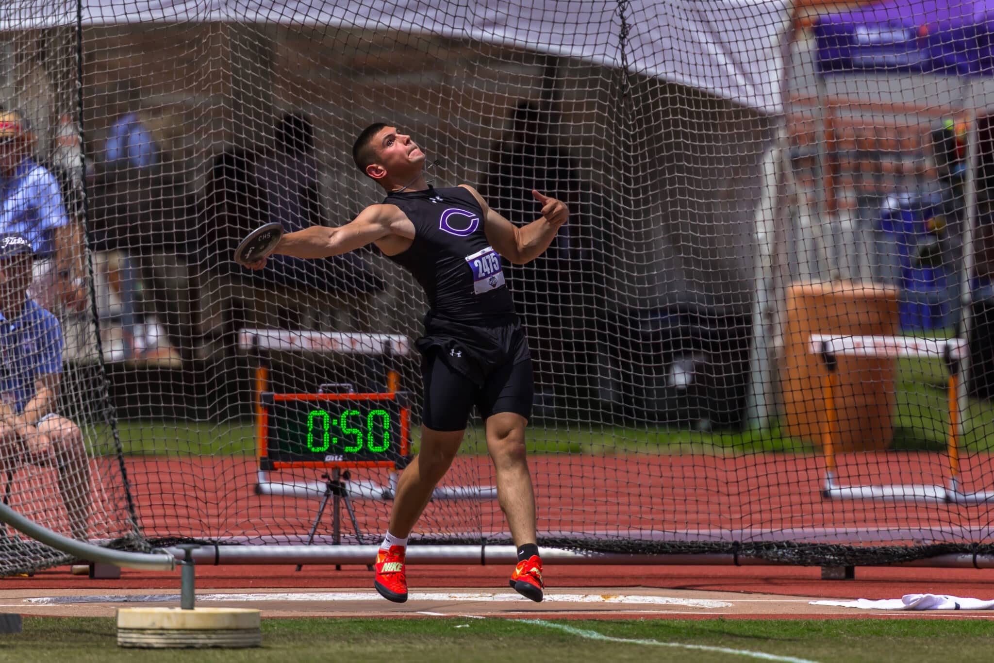 Male discus thrower mid-action inside a throwing net, wearing black athletic gear and red shoes, timer reading 0:50 in the foreground.
