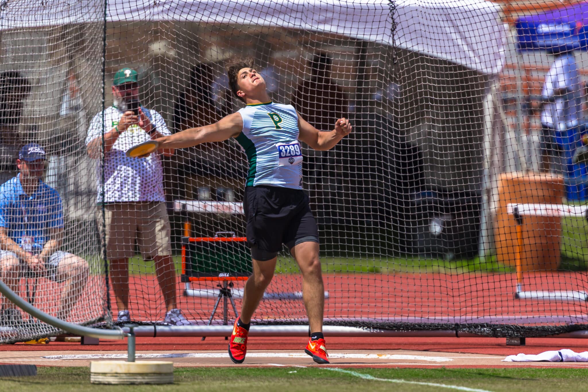 Male discus thrower in a throwing cage mid-action, right arm extended with discus, bib 3209 on chest