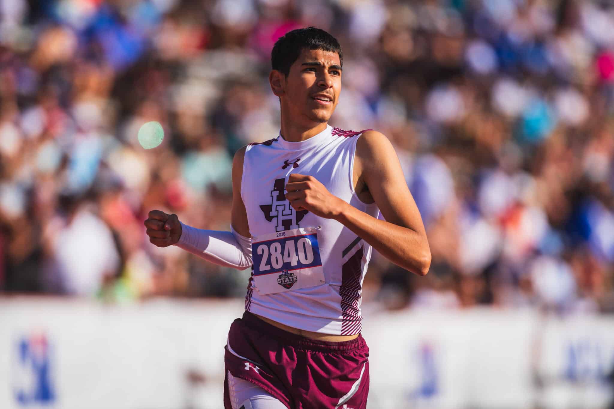 Male sprinter in a white and maroon athletic kit, bib 2840, running with a crowd blurred in the background.