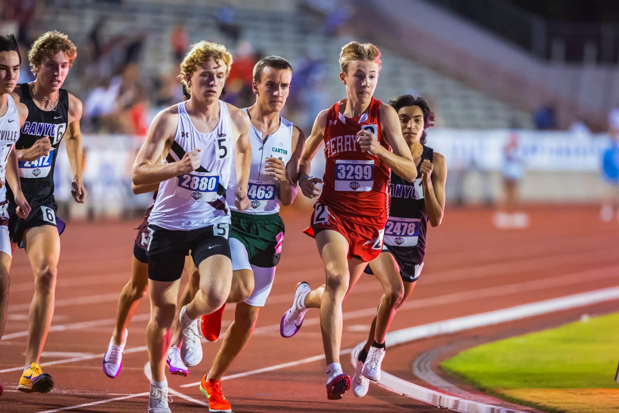 Group of sprinters on a stadium track mid-race, wearing race bibs and colorful uniforms.
