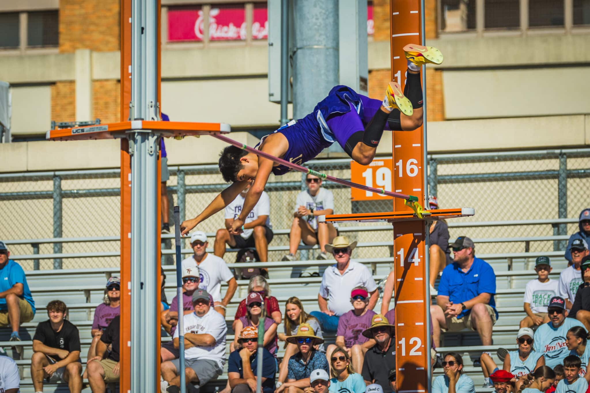 Male pole vaulter in purple uniform clears a high bar during a track and field meet, with spectators in the stands watching.