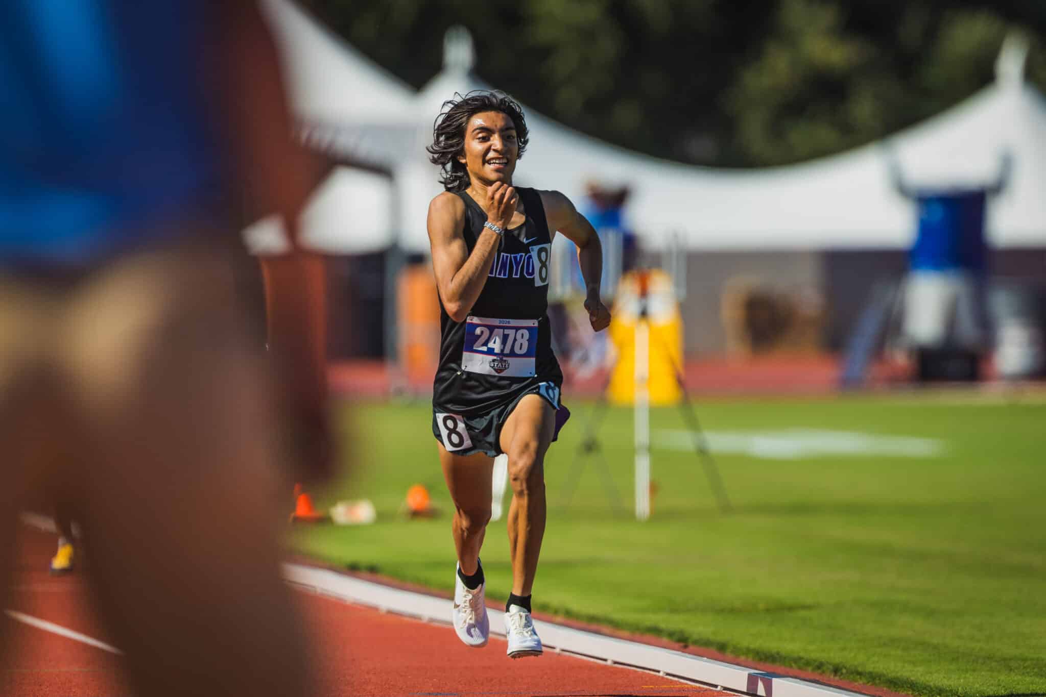 Runner in a black outfit sprinting on a red track during a race, bib number 2478, with blurred spectators and tents in the background.
