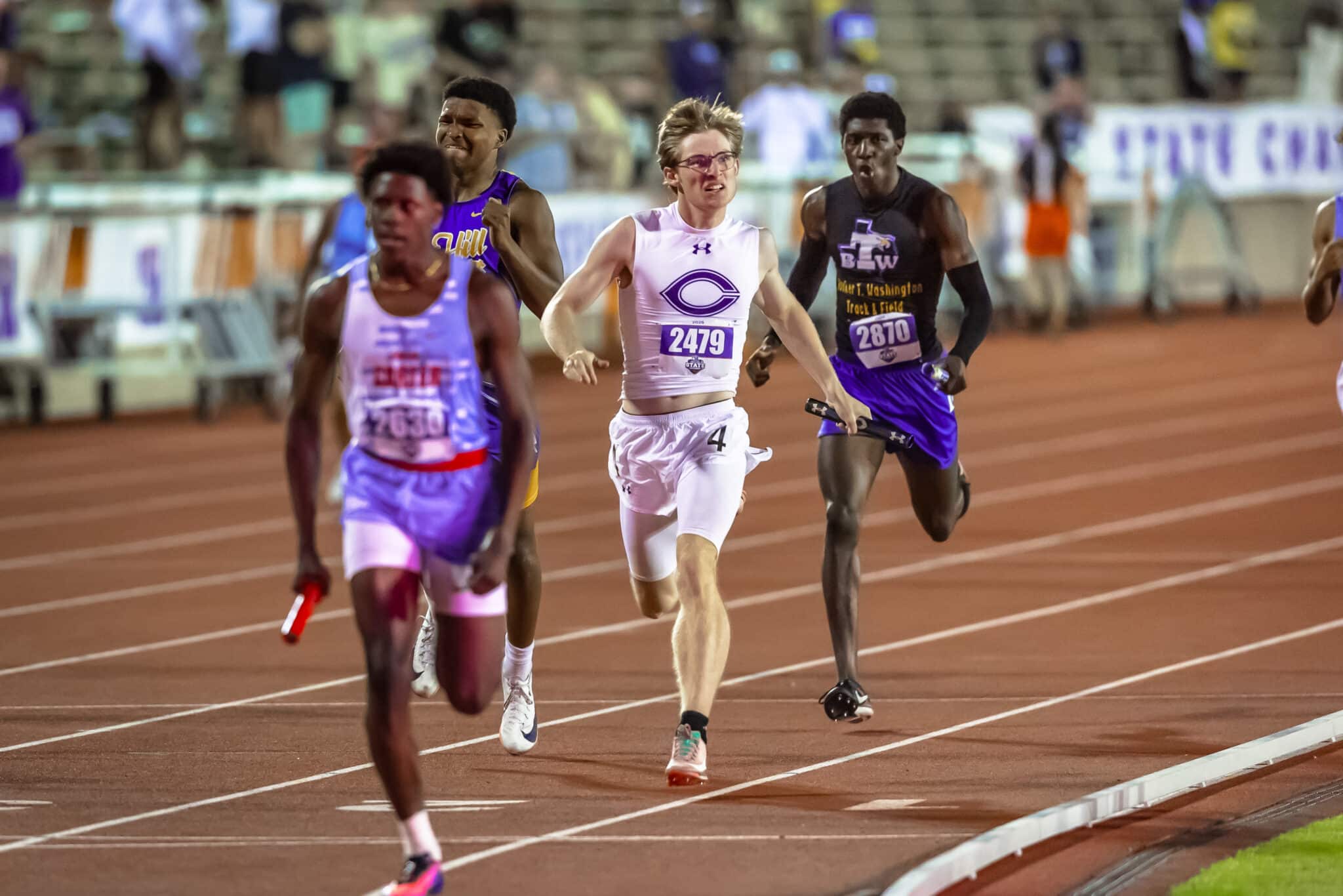 Male sprinters race on a red track; central runner in white with bib 2479 leads the pack.