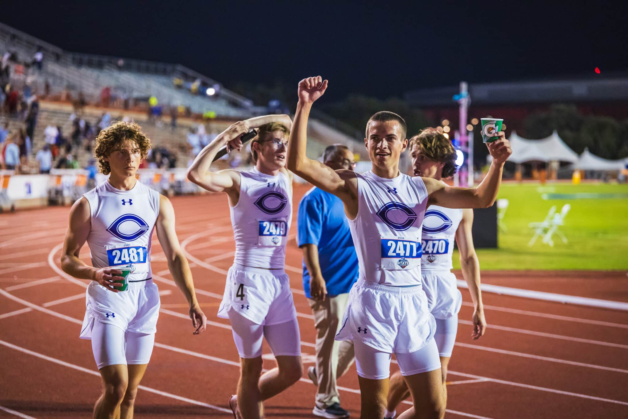 Group of male track runners in white uniforms celebrate on a nighttime stadium track, holding cups of drinks as they finish a race.