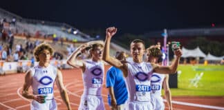 Group of male track runners in white uniforms celebrate on a nighttime stadium track, holding cups of drinks as they finish a race.