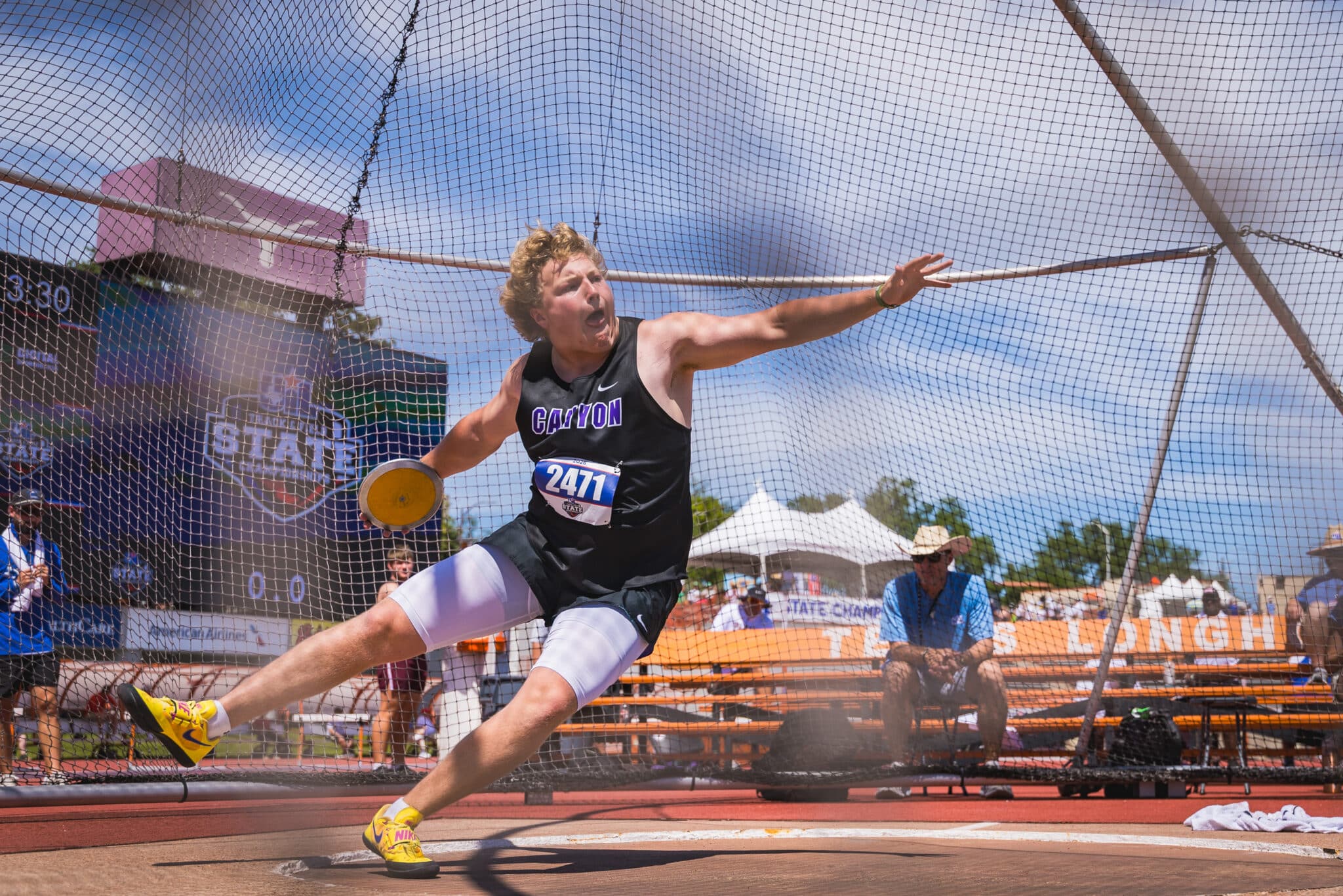 Male track and field athlete in a black Canyon jersey mid-throw inside a nets enclosure, bib 2471, yellow shoes.