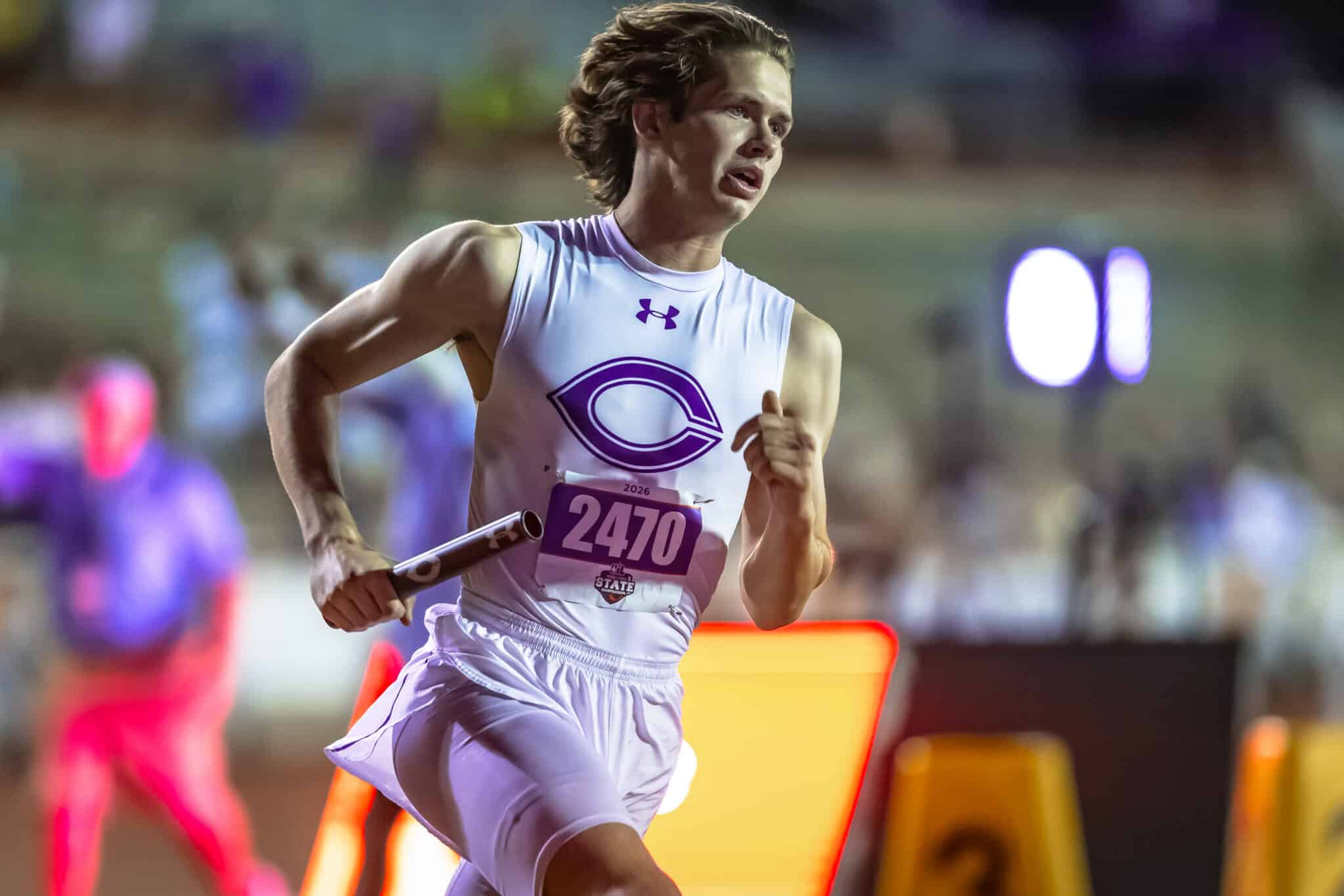 Male sprinter in a white singlet with purple logo, bib 2470, sprinting in a relay race while gripping a baton against a blurred stadium background.
