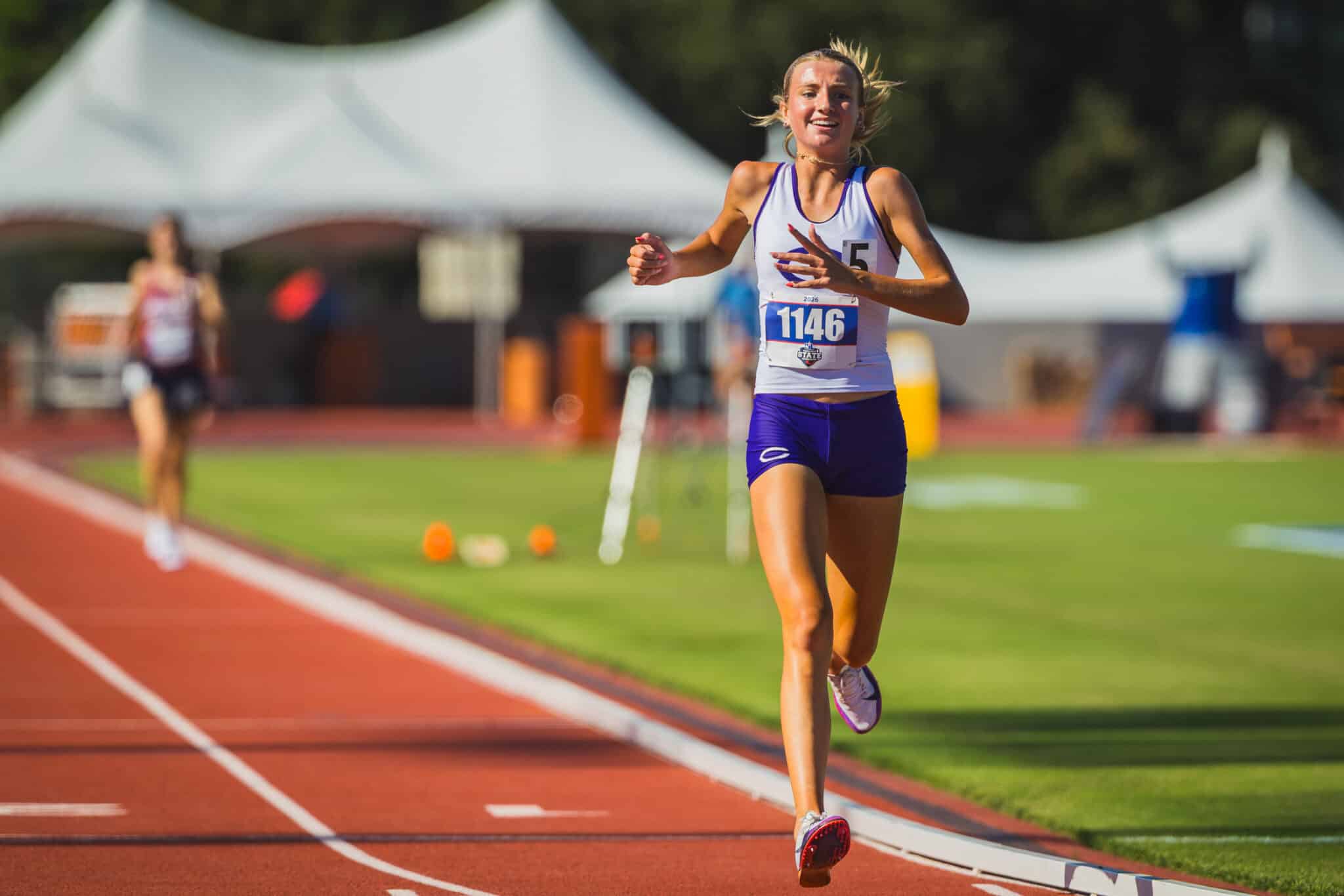 Female runner wearing bib 1146 on a red track, mid-race with blurred stadium tents behind.