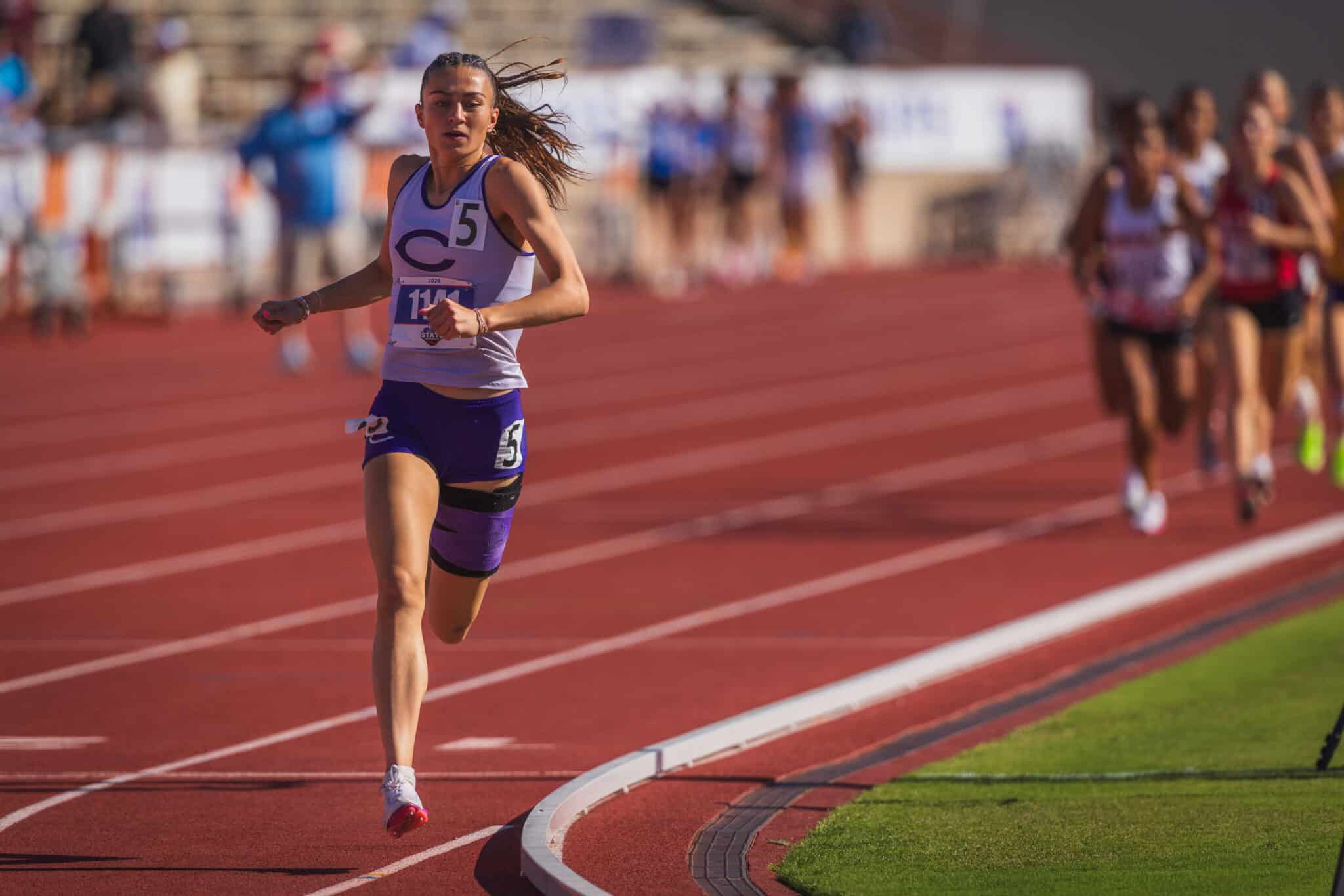 Female sprinter in purple uniform runs on a red track, bib number 5 visible, mid-race on curve with blurred spectators in the background, outdoor stadium setting.