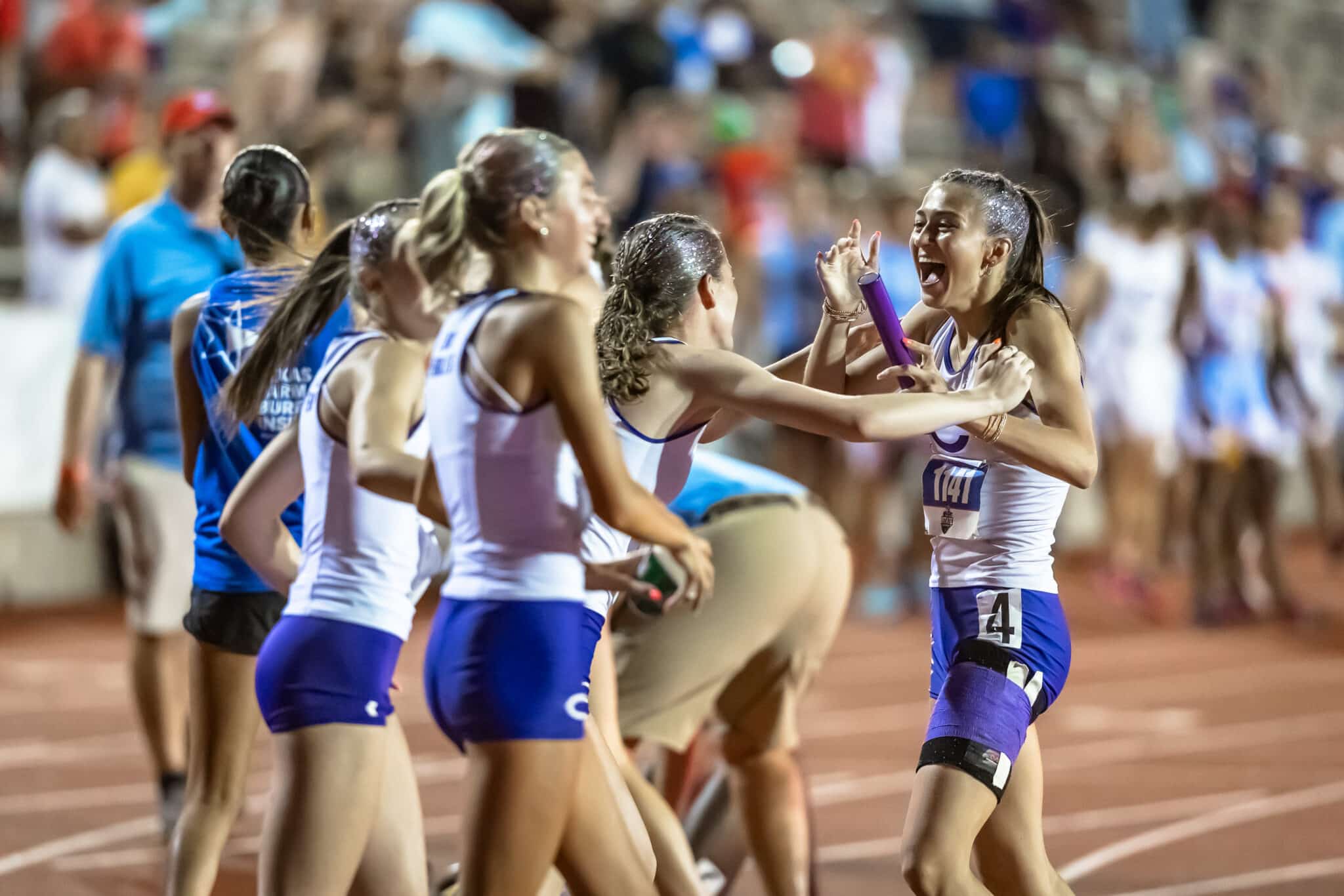 Female runners in white and blue uniforms celebrate on a track, high-fiving and smiling as a crowd cheers in the background, with a purple baton visible among them.