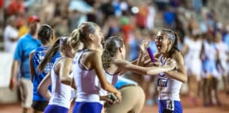 Female runners in white and blue uniforms celebrate on a track, high-fiving and smiling as a crowd cheers in the background, with a purple baton visible among them.