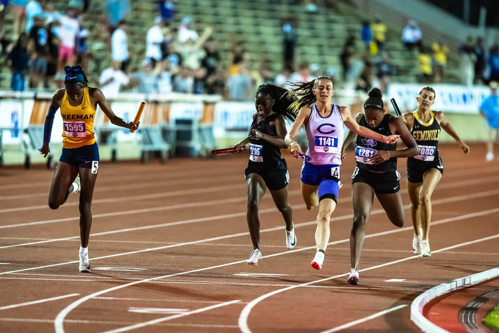 Group of female sprinters in a relay race sprinting on a red track, baton exchanged on the left, spectators blurred in the stands behind them.