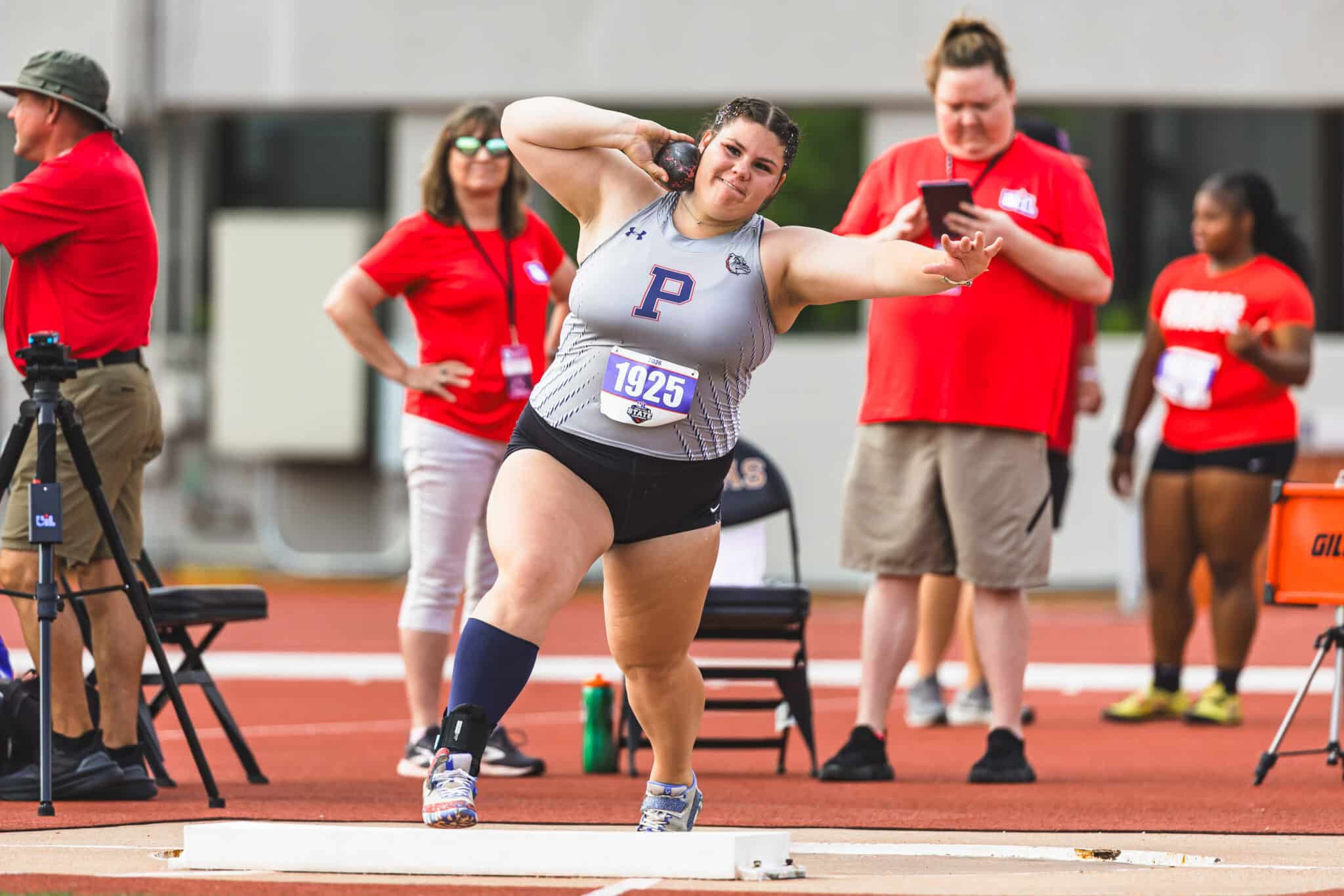 Female athlete in a gray singlet and black shorts mid‑throw of a shot put, bib 1925, on a red track with onlookers in red shirts behind.”,