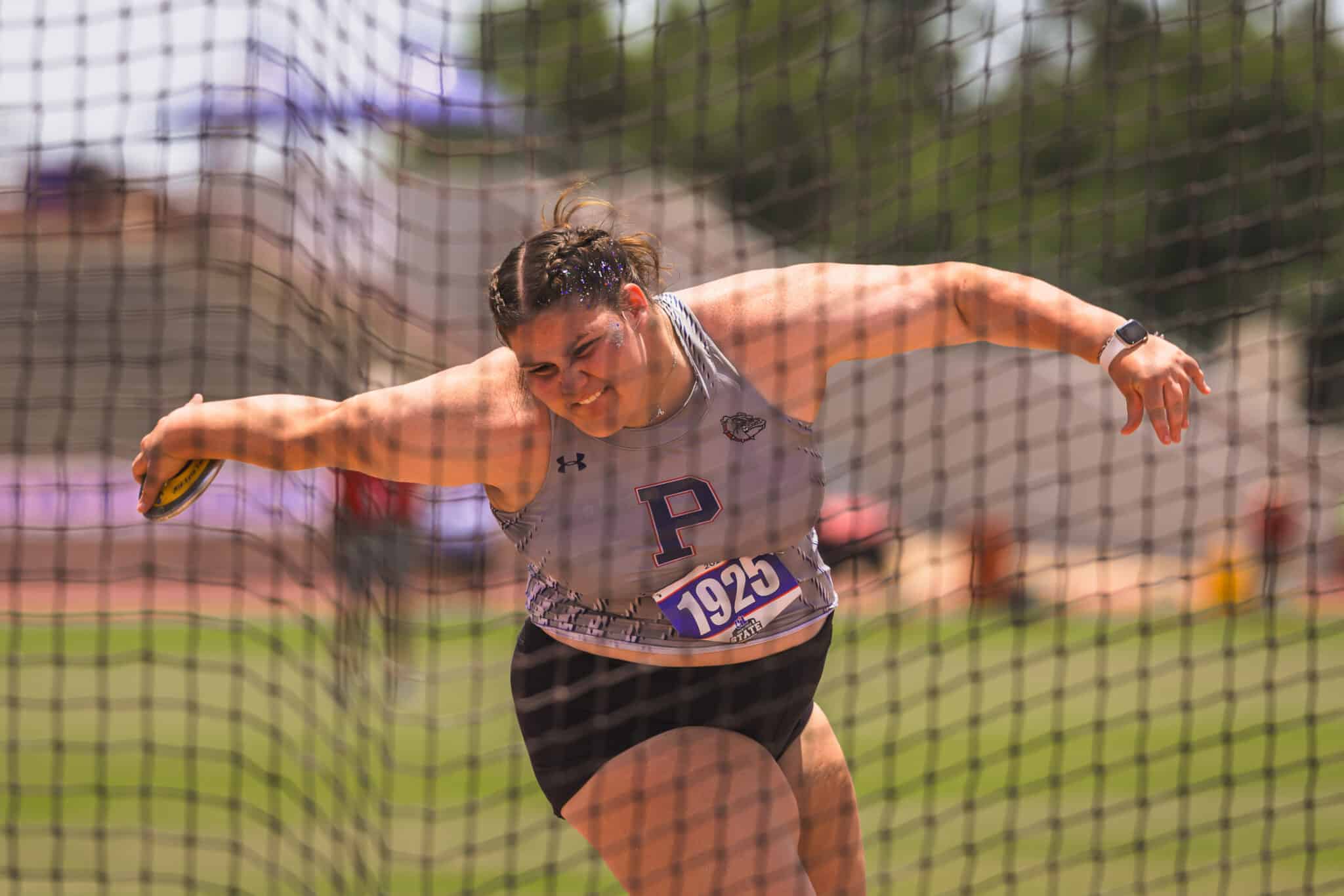 Female track athlete sprinting on a track, wearing a gray uniform with bib 1925, viewed through a protective net.