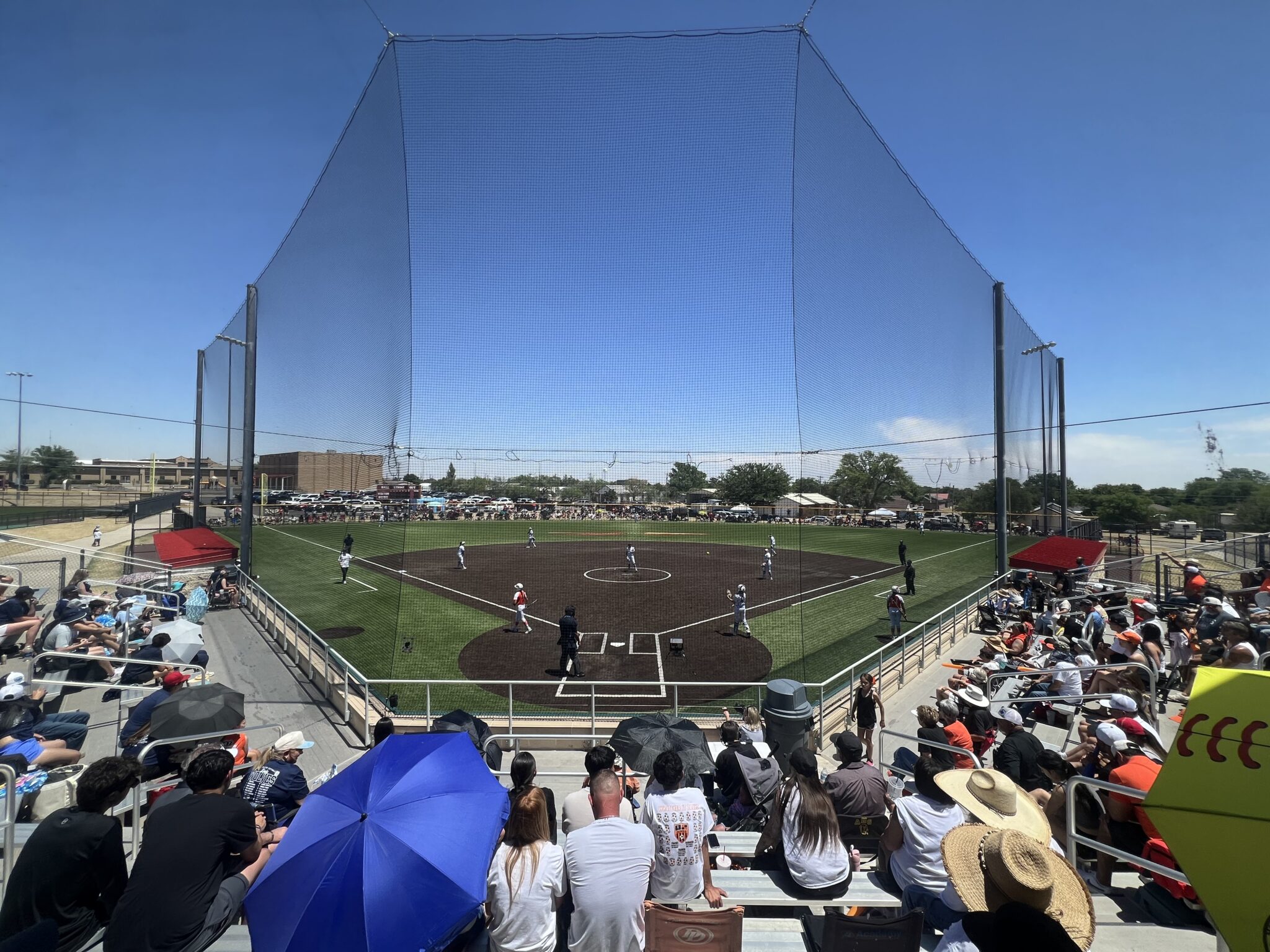 Baseball field in a stadium viewed from the stands with protective netting and spectators seated on both sides under blue sky