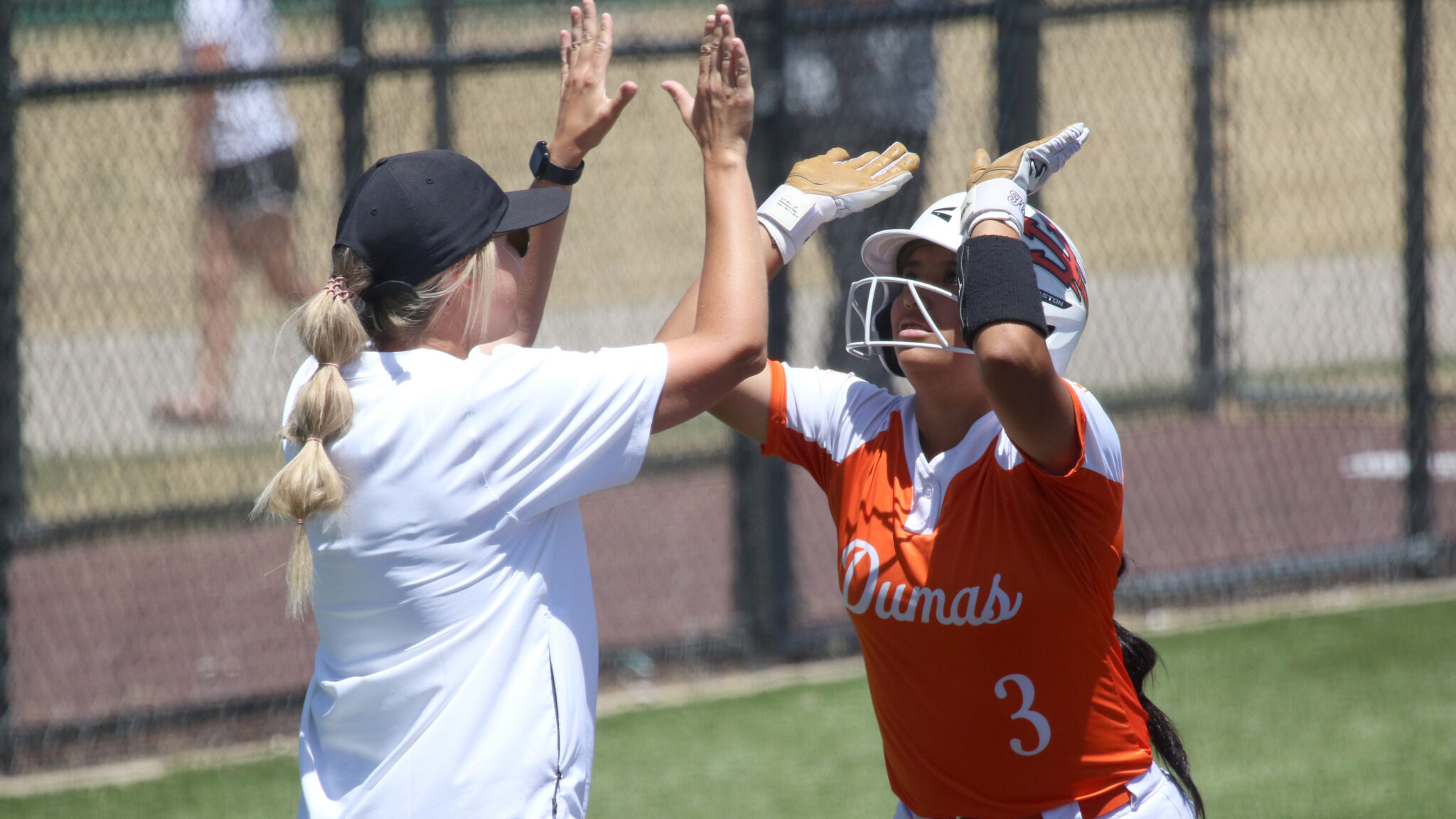 Two softball players high-fiving on a sunny field; one wearing an orange jersey and helmet, the other in a white shirt and cap.