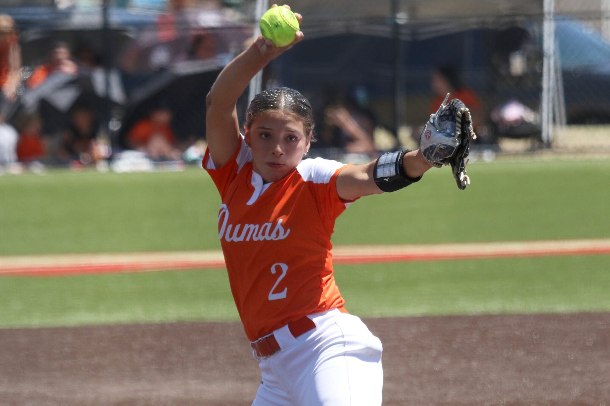 Female softball pitcher in an orange jersey throws a neon green ball, glove up, on a grassy field.