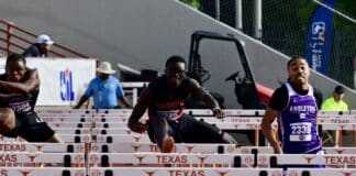 Male sprinter in black uniform clears a hurdle mid-race at a Texas track meet, with other runners nearby.