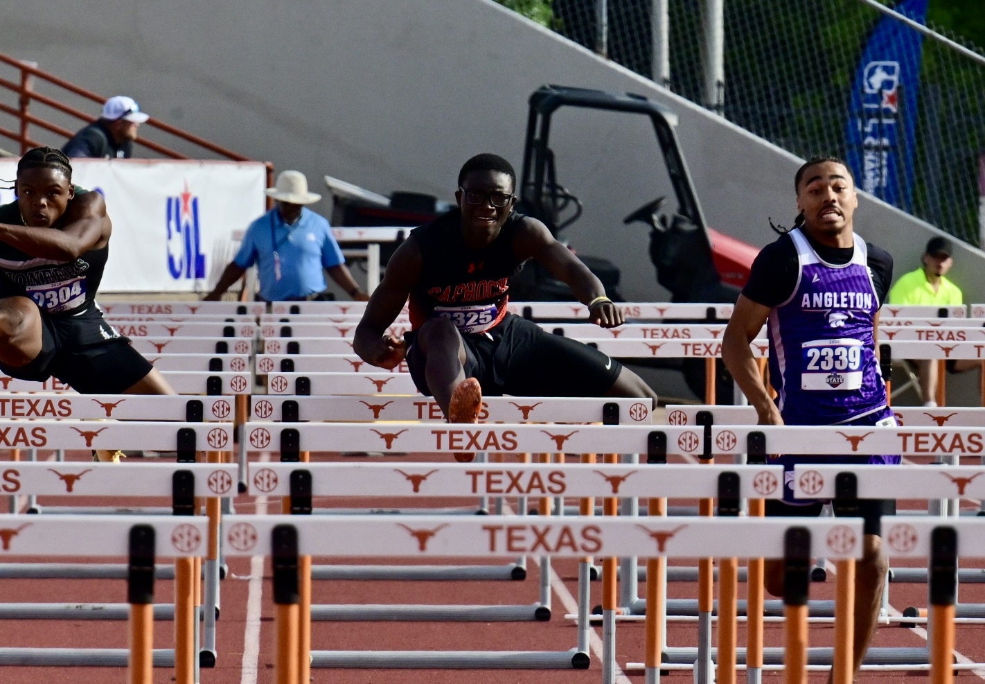 Male sprinter in black uniform clears a hurdle mid-race at a Texas track meet, with other runners nearby.